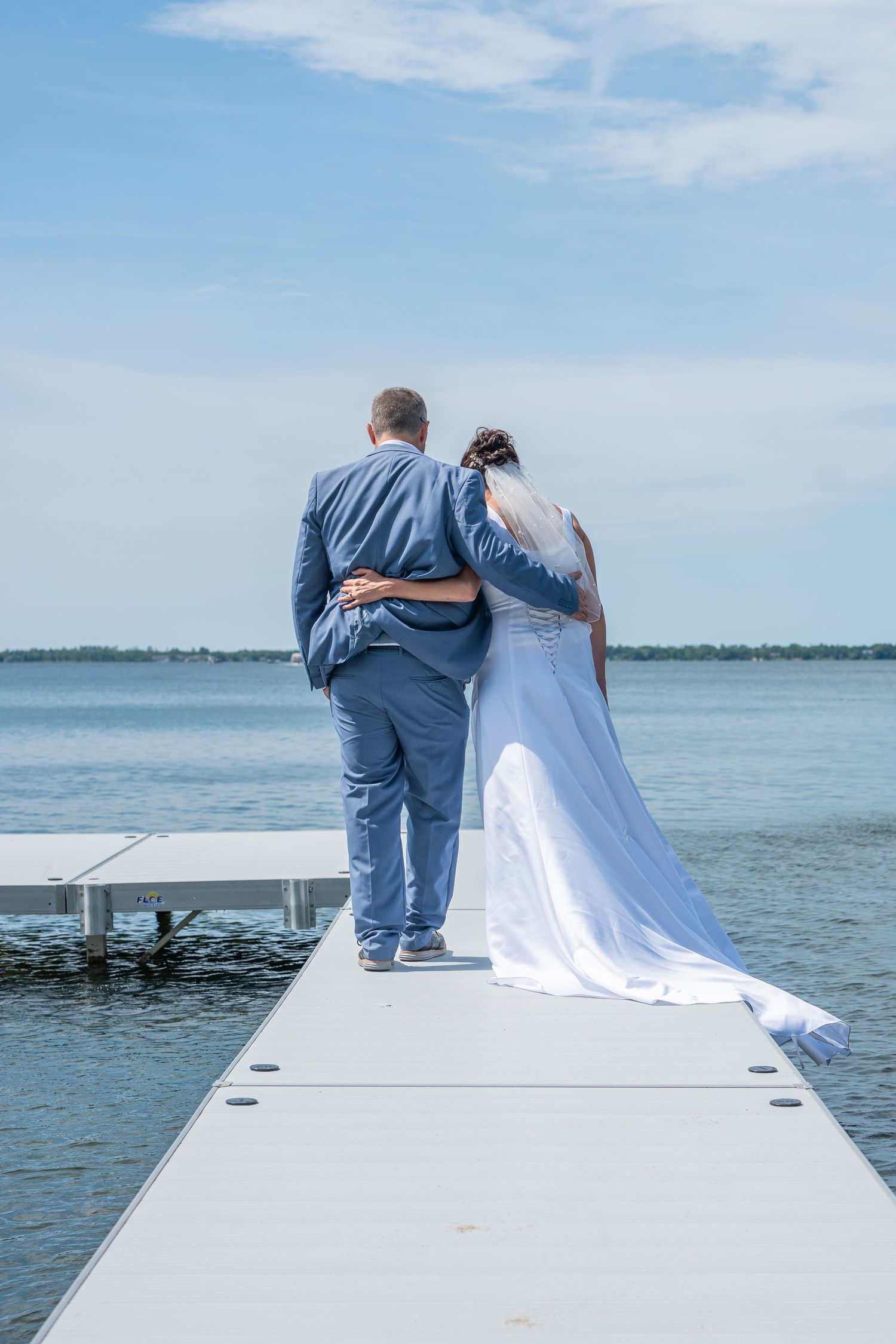 Wedding couple embraces while sitting on edge of white dock with calm water and blue sky behind them.