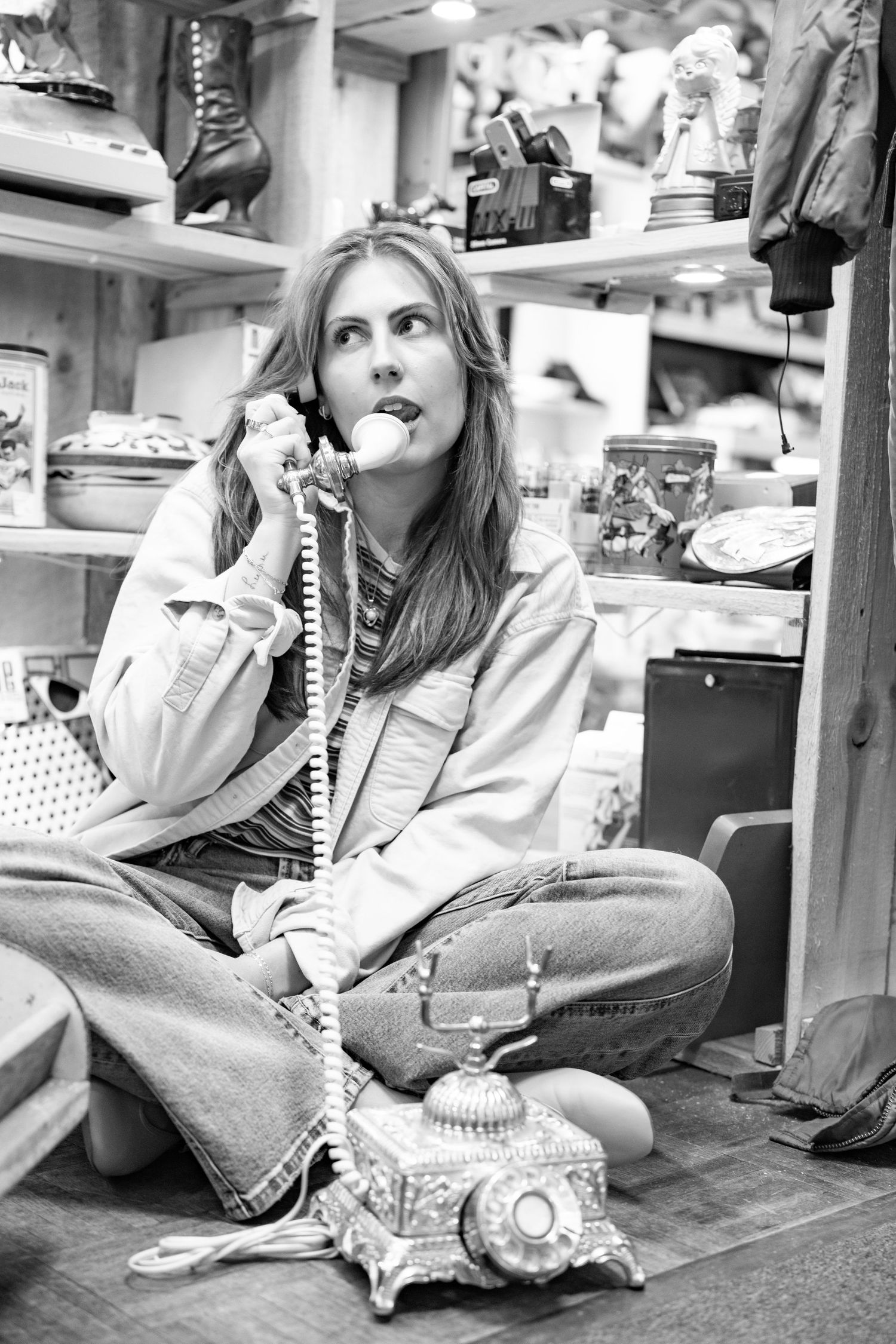Black and white photo of a woman sitting cross-legged on the floor, talking on a vintage ornate telephone in a antique shop.