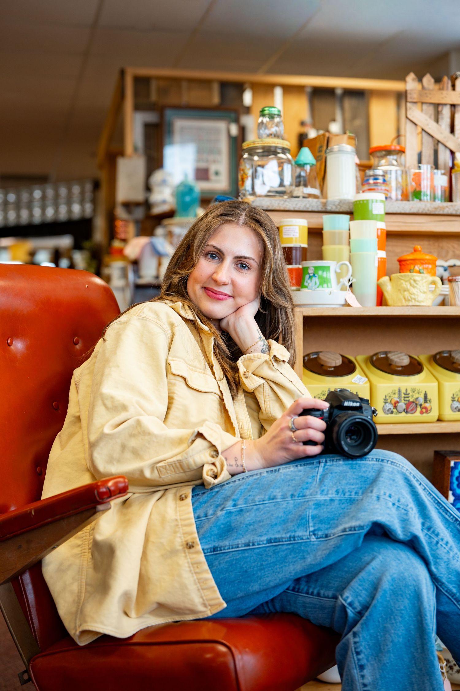 Woman sitting in red chair holding Nikon camera, smiling in vintage shop with colorful retro items on shelves behind her.