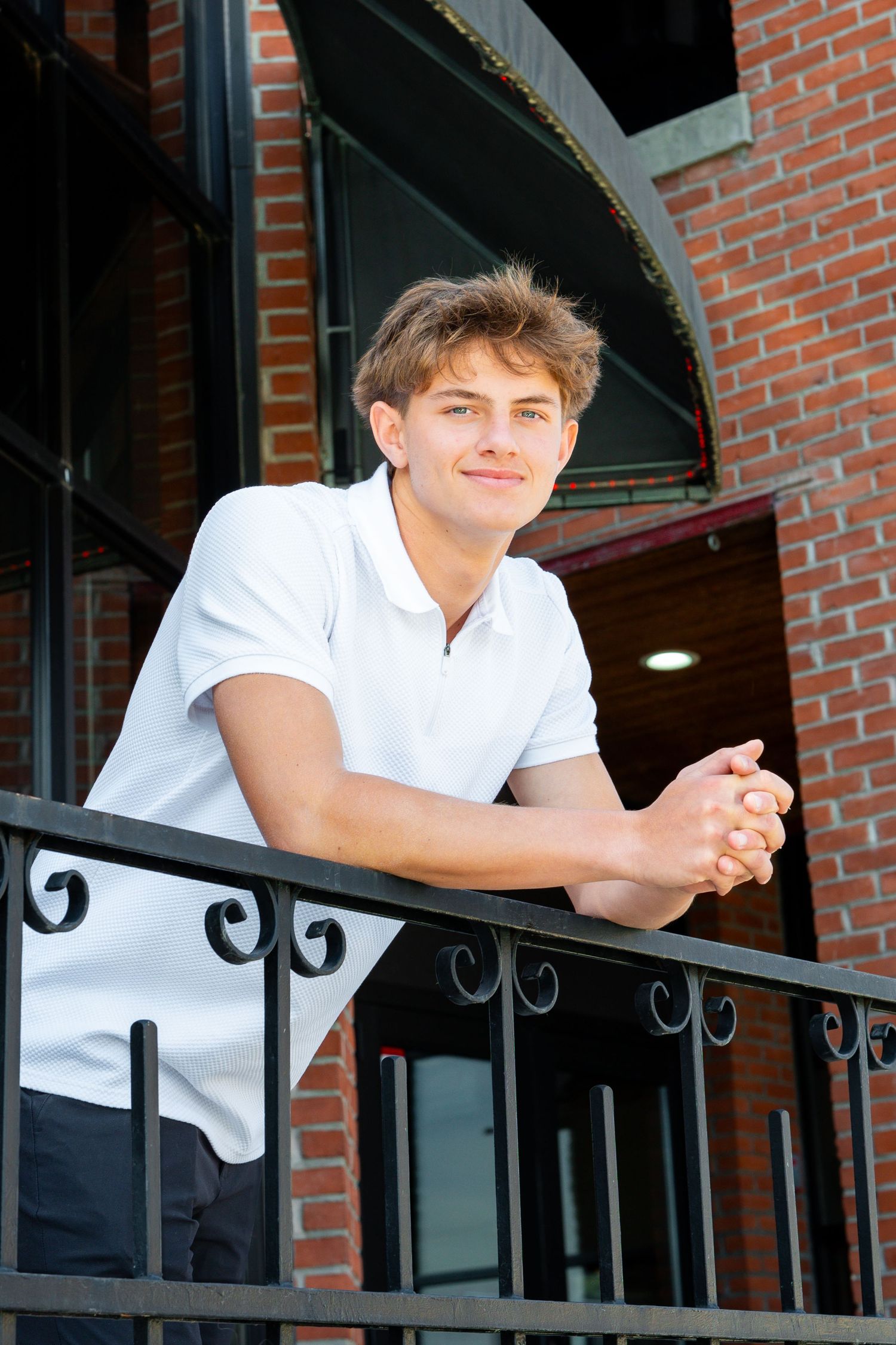 A young person in a white polo shirt leans against a black iron railing on a brick building balcony.