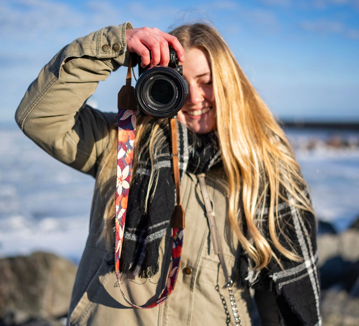 Smiling blonde woman photographer holding a DSLR camera outdoors in winter, wearing a plaid scarf and olive jacket.