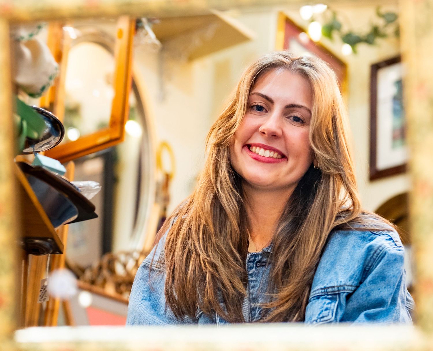 Smiling young woman with long blonde hair wearing a denim jacket, posing in a decorative shop interior.