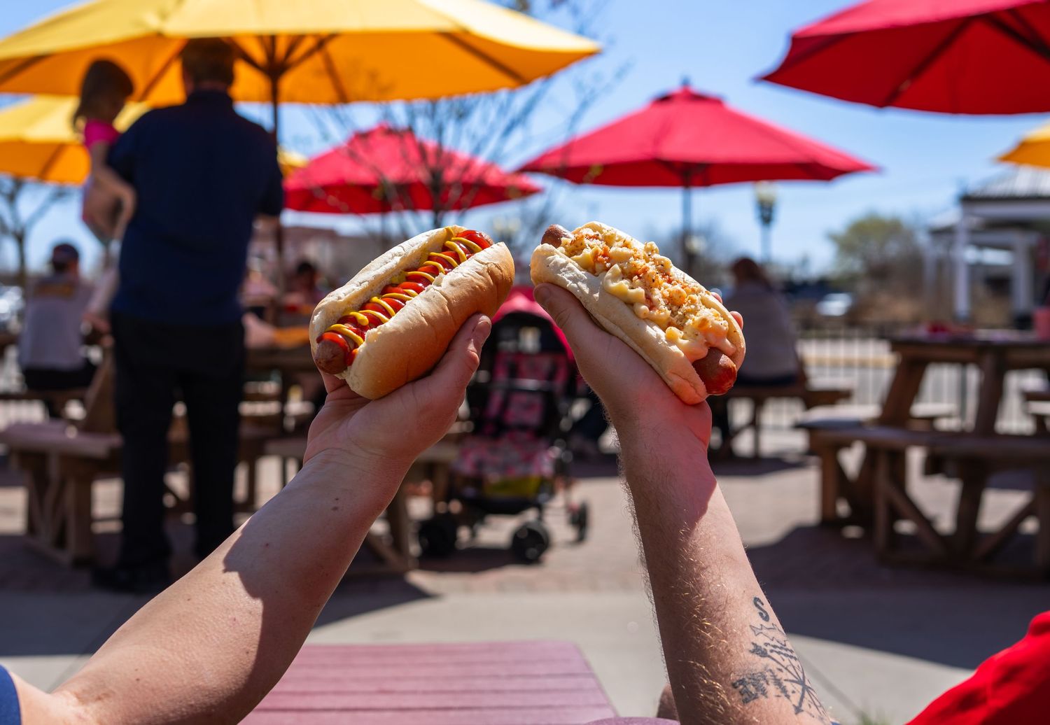 Hot dogs being held up against a sunny outdoor backdrop with colorful umbrellas.