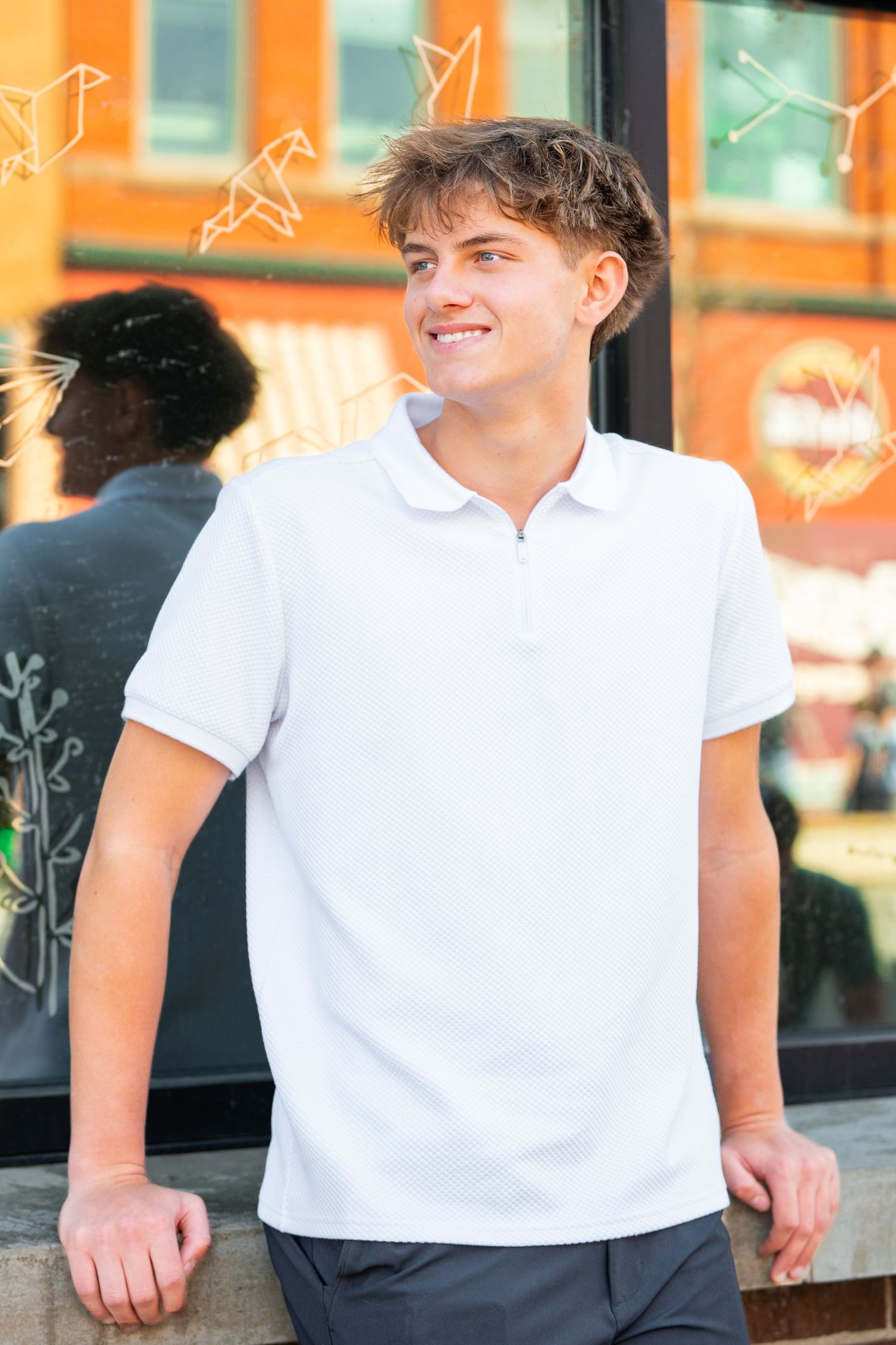 A person in a white polo shirt and dark pants stands against a blurred orange and white background.