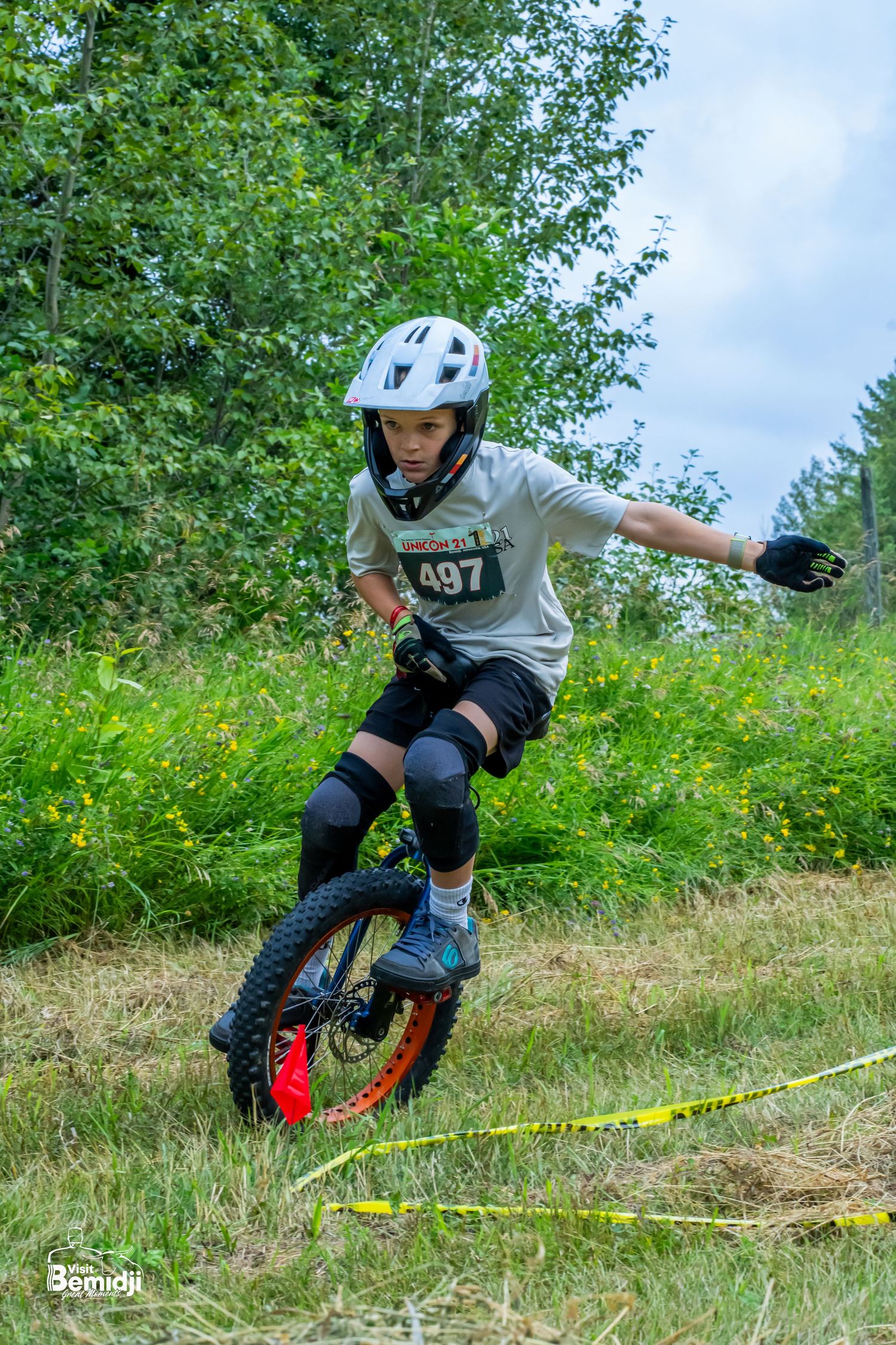 Unicyclist jumping over obstacles on a grassy dirt course wearing protective gear.