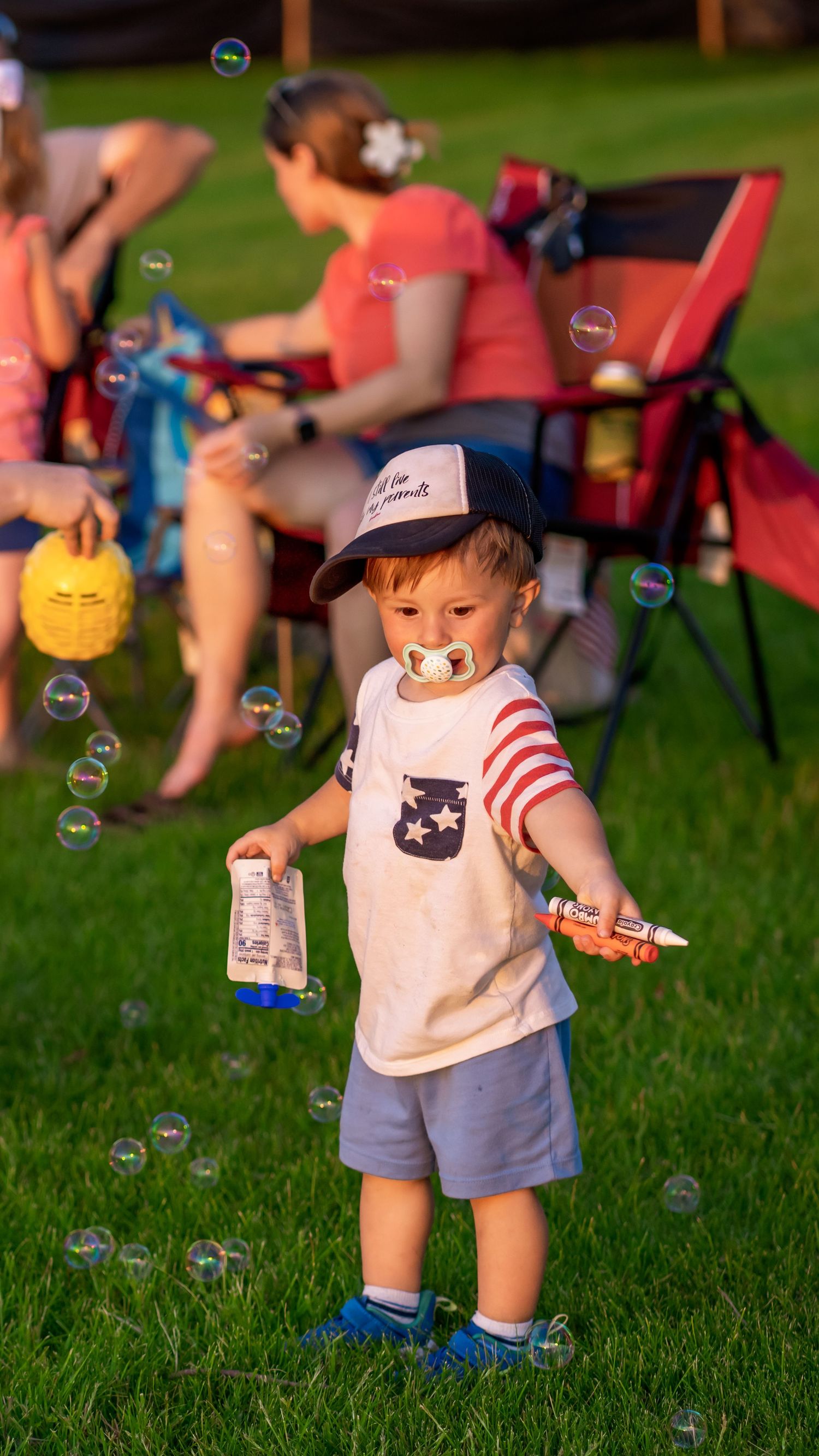 Small bubbles float through the air as a toddler plays on grass during a summer gathering.