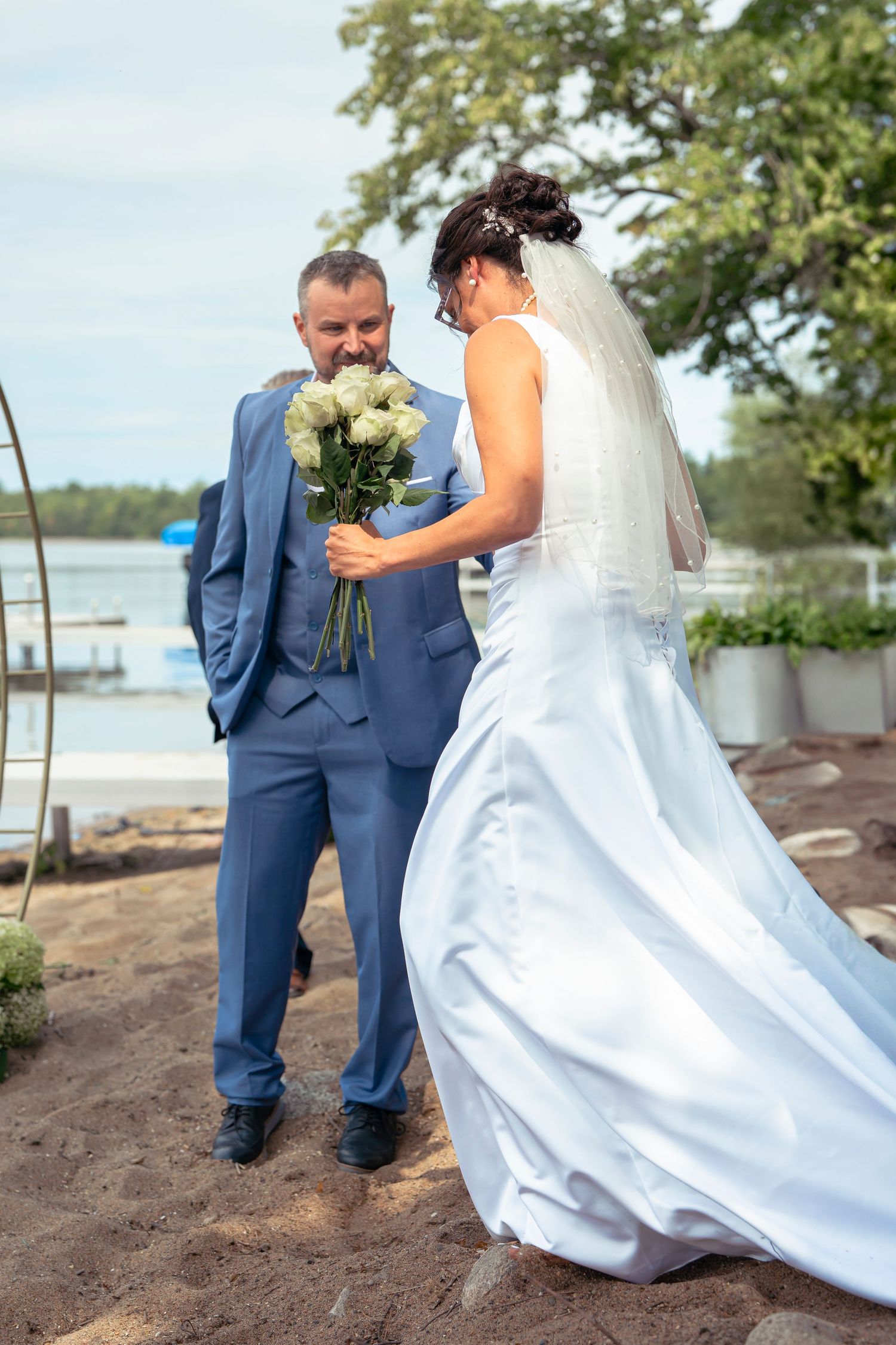 Wedding couple walking along sandy beach with water and boats in background.