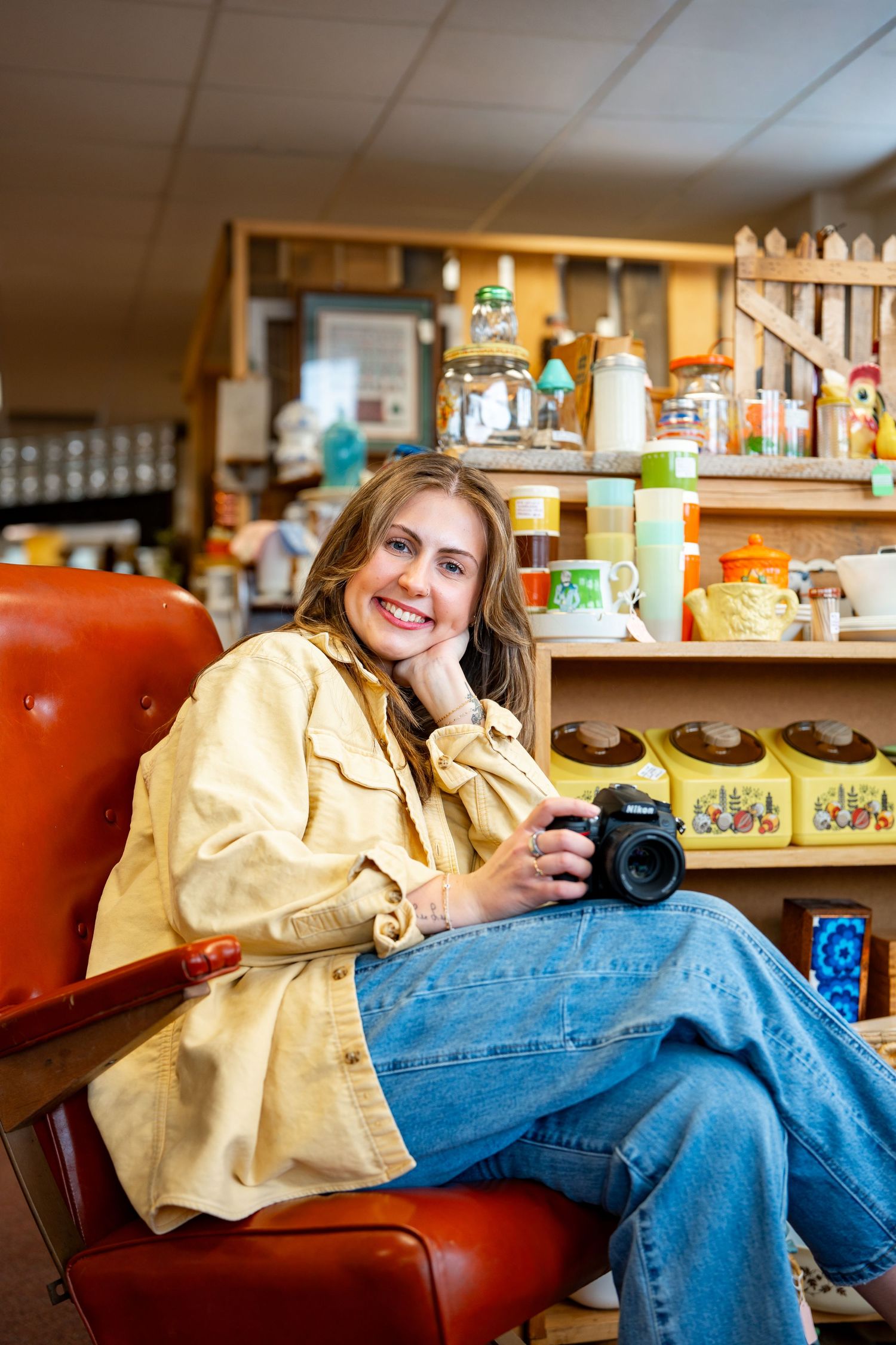 Smiling woman sitting in red chair holding DSLR camera in a vintage shop with colorful collectibles on shelves behind her.