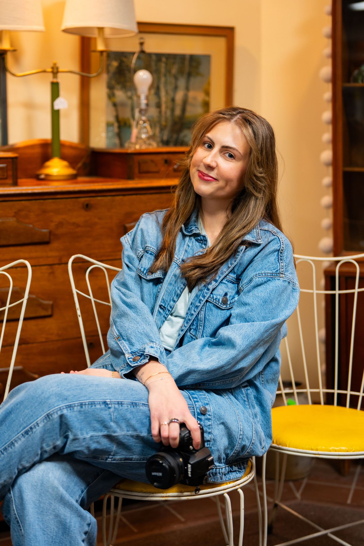 Young woman in denim jacket sitting on chair holding camera in vintage-styled room with wooden dresser.