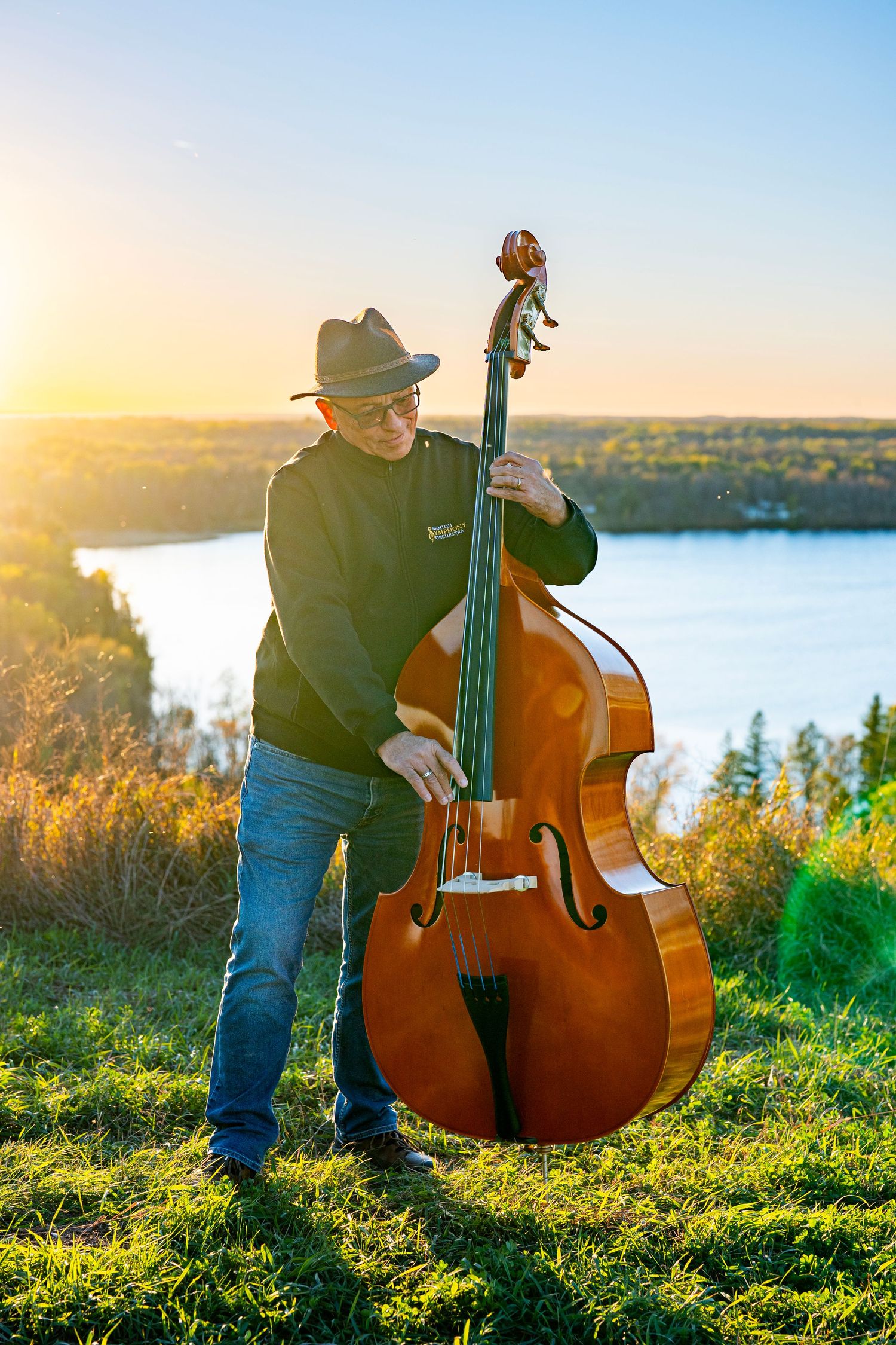 Upright bass player performing on a hilltop overlooking a lake at sunset.
