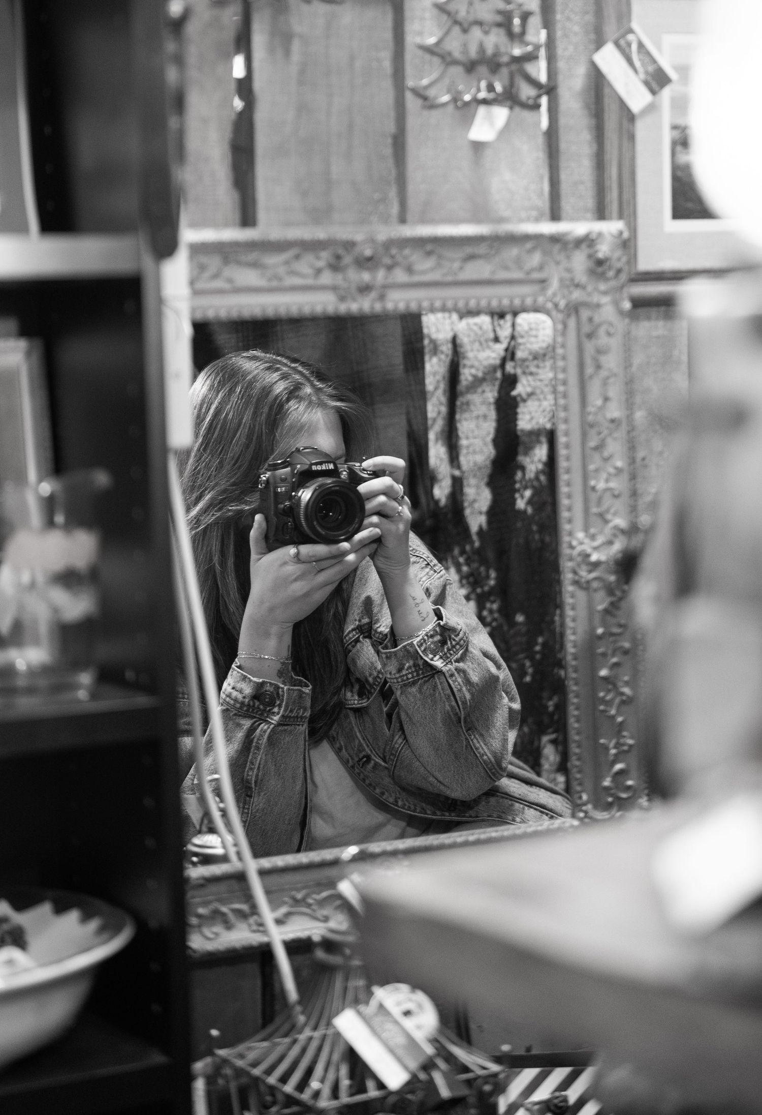 Black and white photo of a woman taking a mirror selfie with a film camera in a vintage shop.