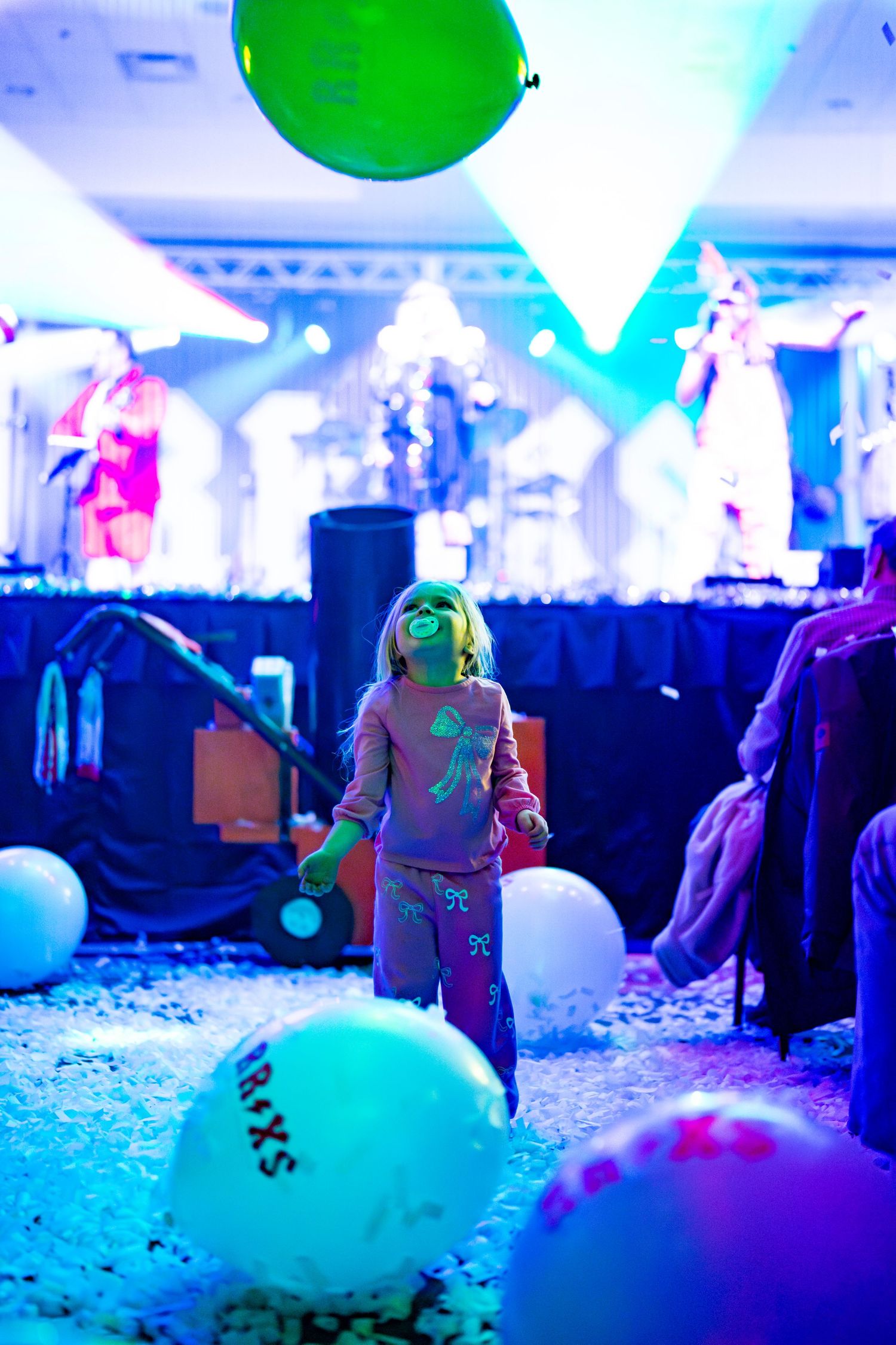 Colorful concert scene with green balloons and confetti under blue lighting in an indoor venue.