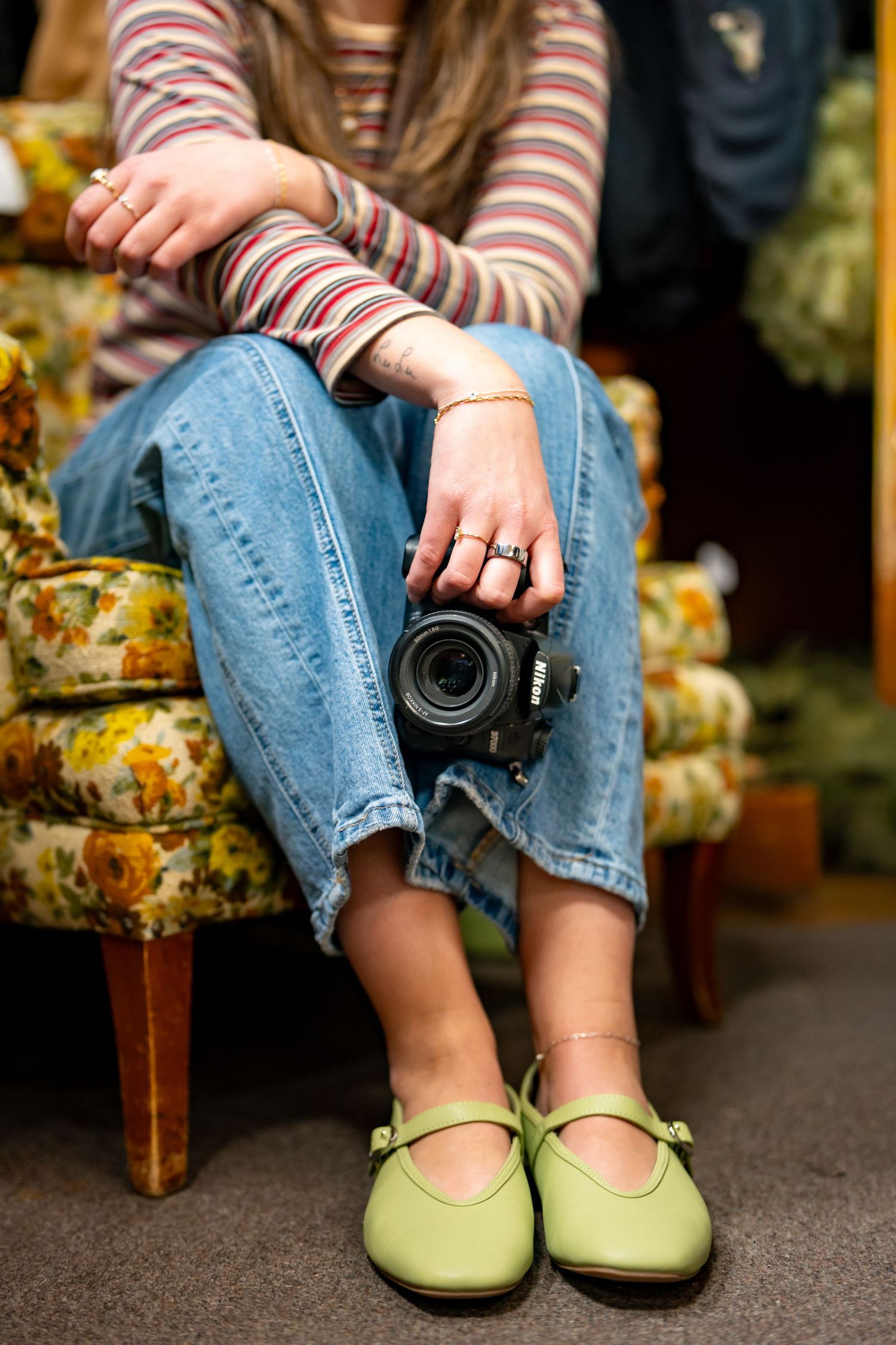 Woman in striped top and jeans holding a Nikon camera, wearing green Mary Jane flats, seated on a floral armchair.