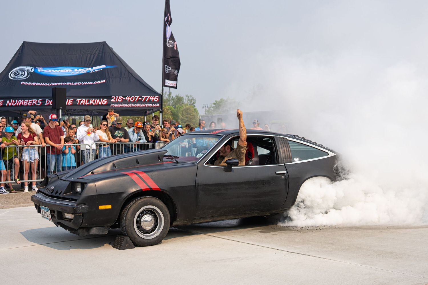 Black muscle car performing a burnout with smoke billowing from rear tires at a car show.