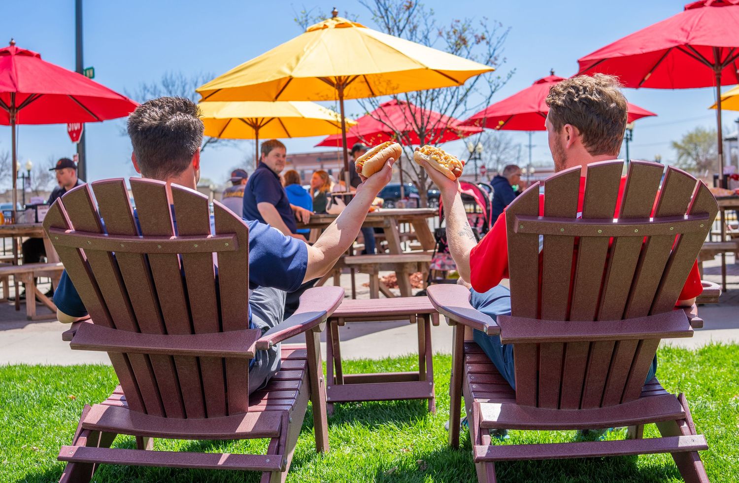 People relaxing in Adirondack chairs under red and orange patio umbrellas with hotdogs.