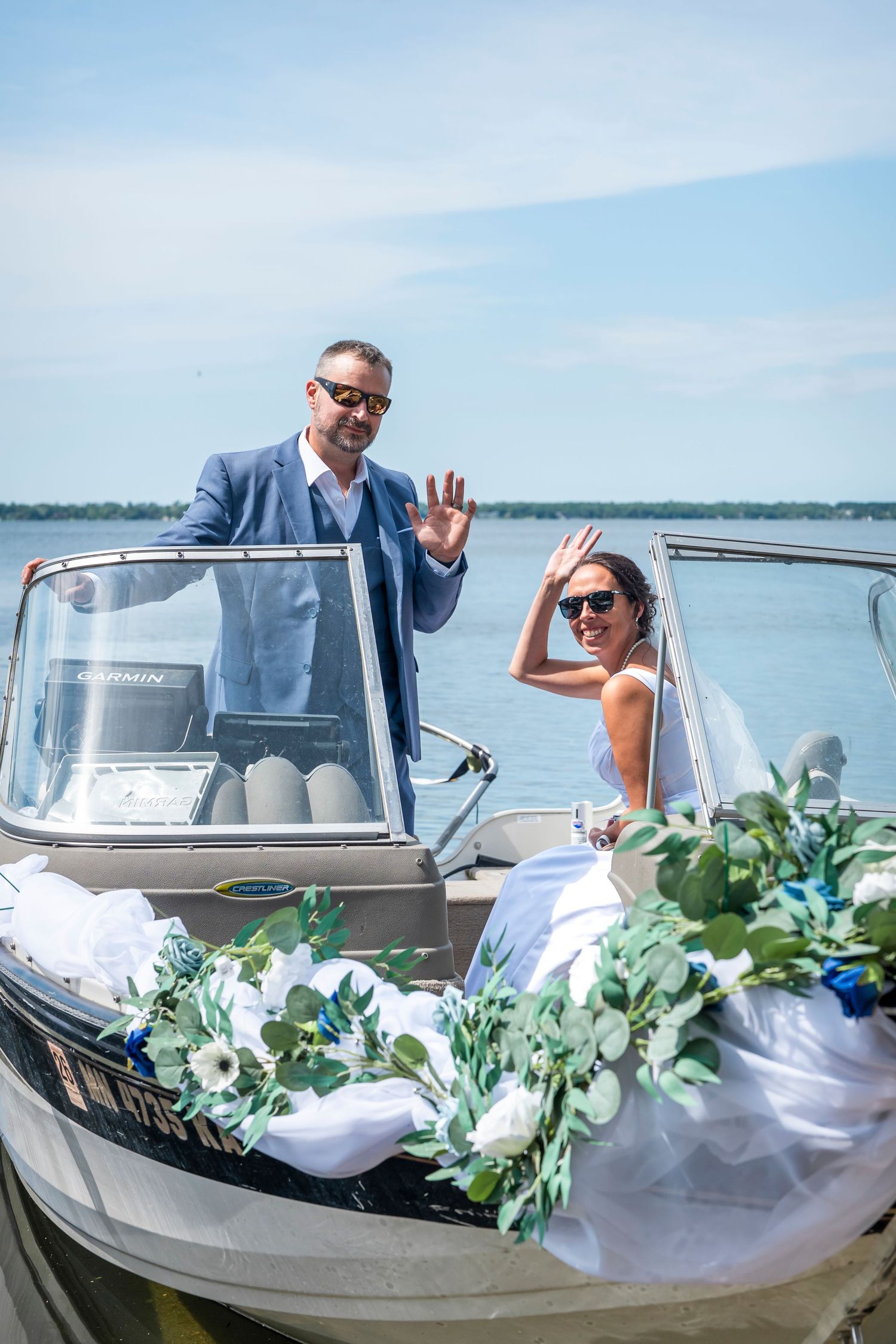 Newlyweds waving from a decorated boat on a sunny lake.