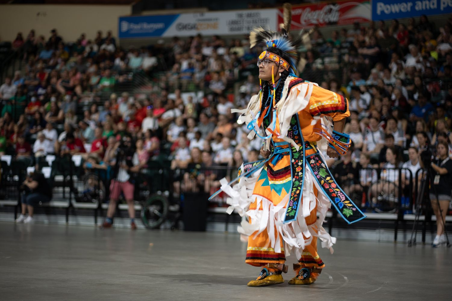 Traditional Native American dancer performs in colorful regalia at indoor event.