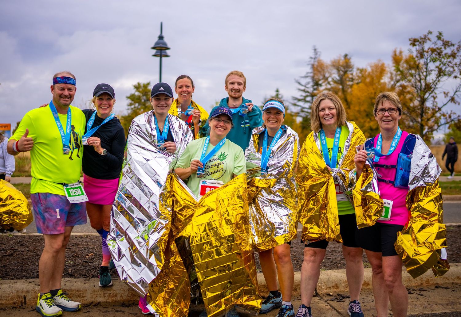 Group of marathon finishers wrapped in gold thermal blankets celebrating outdoors.