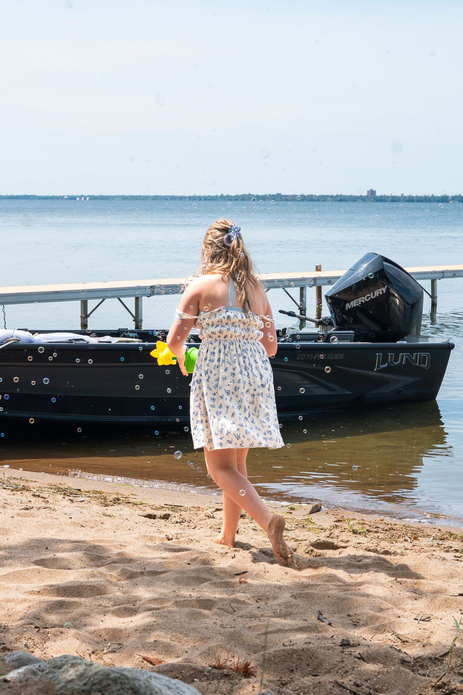 Kid in floral dress stands on beach dock near boat on sunny day playing with bubbles.