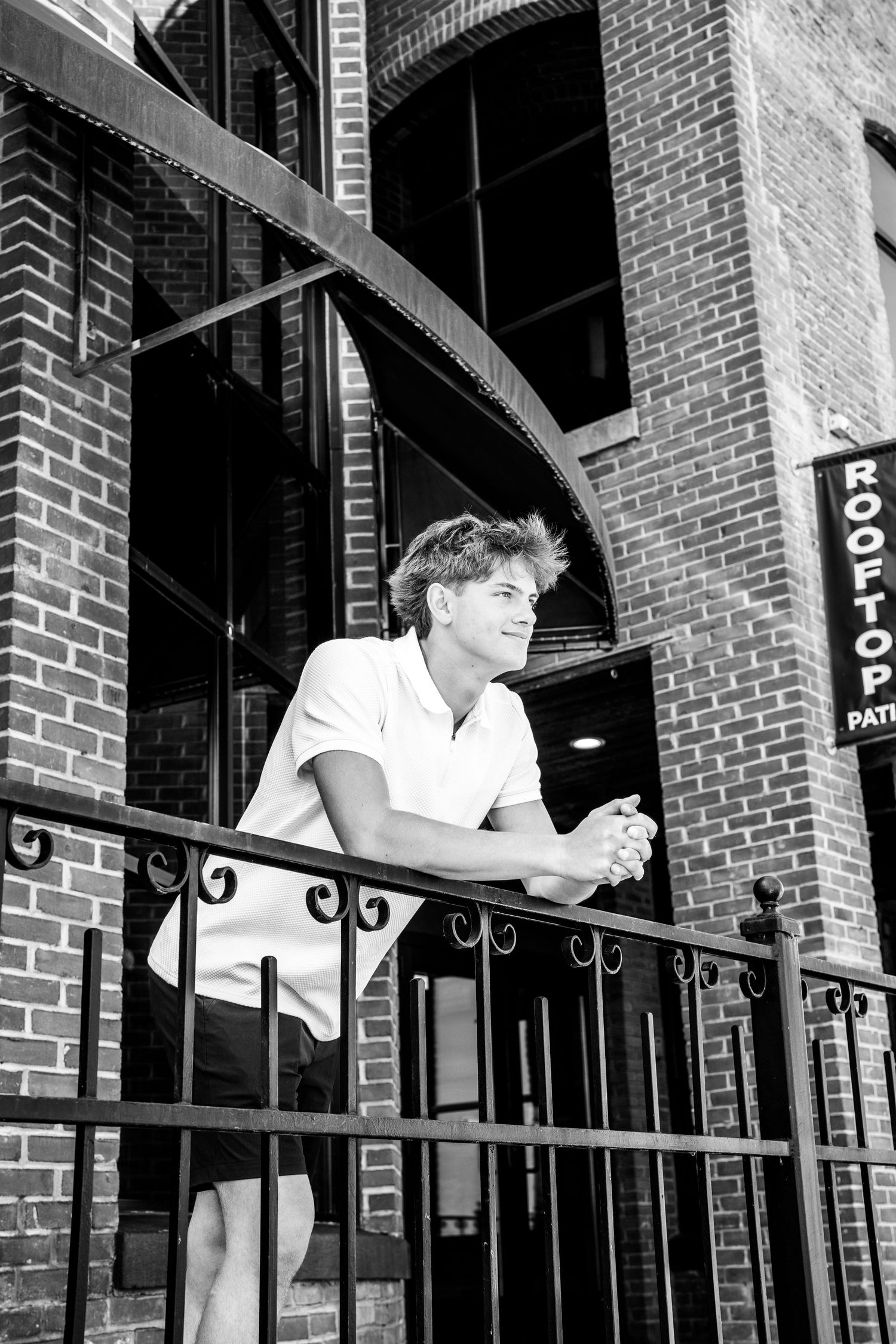 Black and white photo of someone in a white shirt leaning on a wrought iron balcony railing outside a brick building.