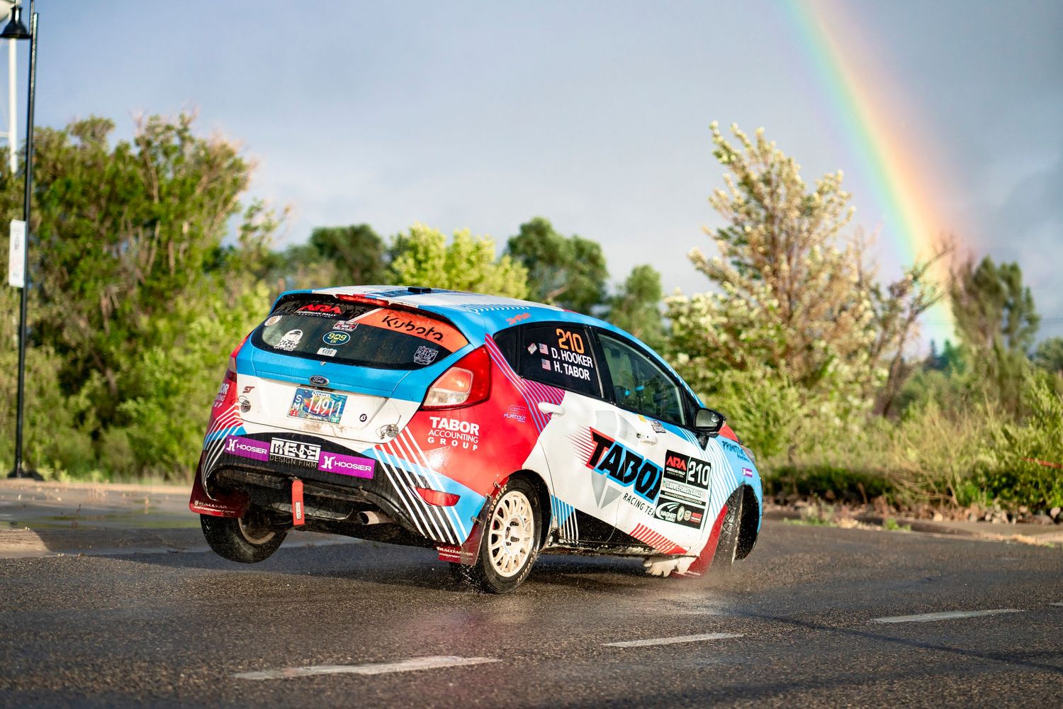 Rally car driving through corner with rainbow visible in background.