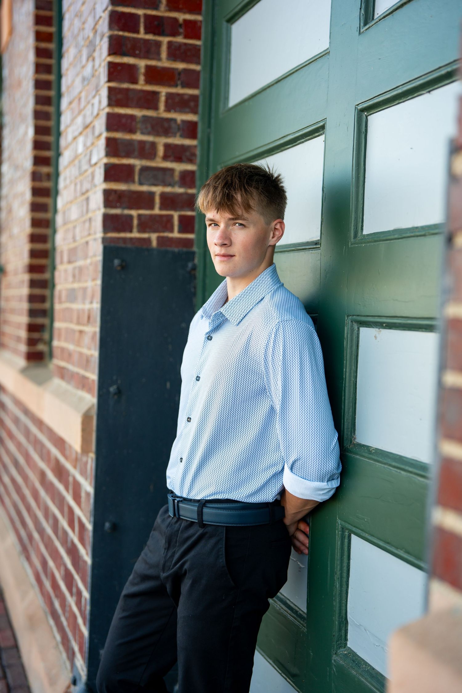 Fashion portraits against a green door and brick wall in light blue shirt and dark pants.