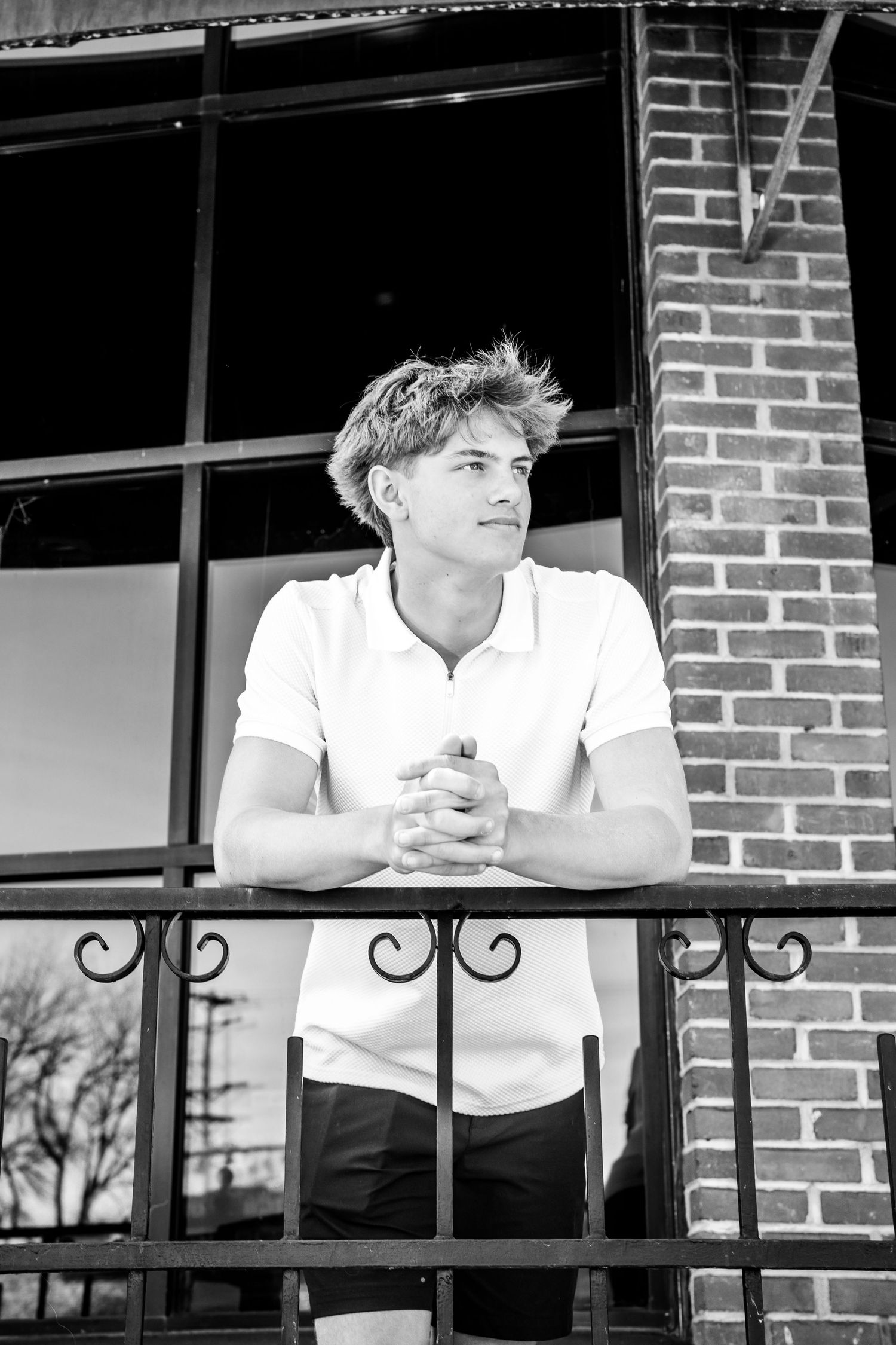 A person in a white shirt leans on a brick building's railing in a black and white photo series.