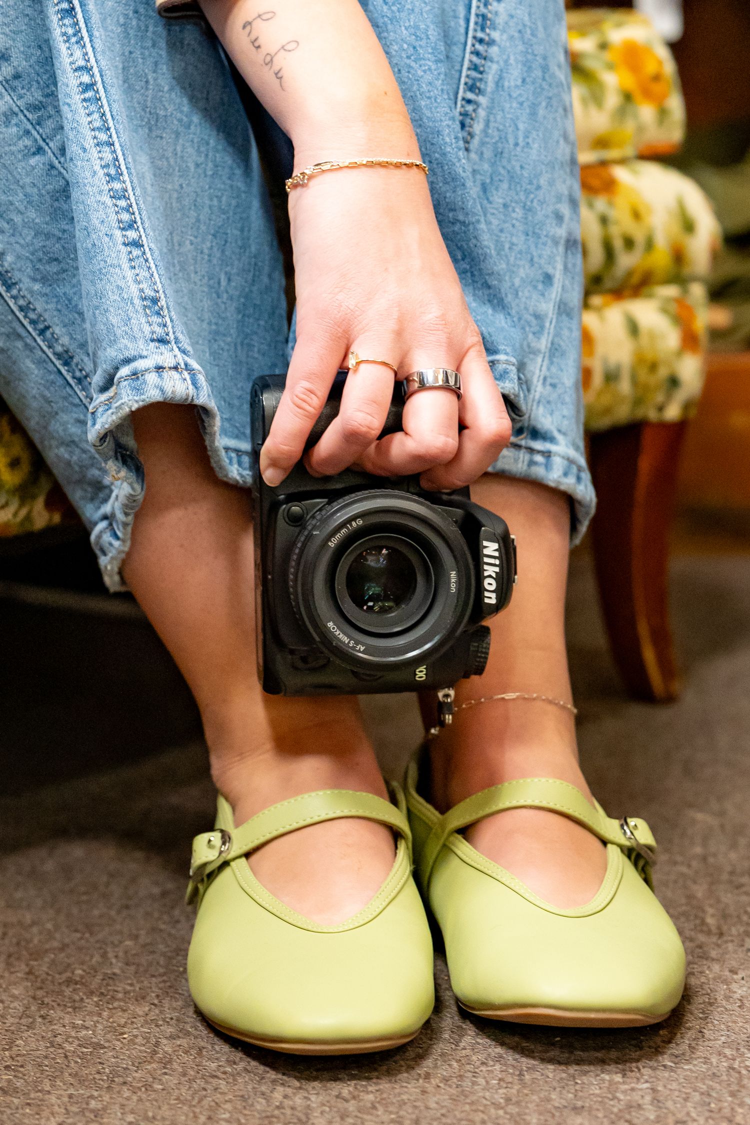 Person sitting in floral chair holding Nikon camera, wearing green Mary Jane flats, jeans, gold rings and bracelets.