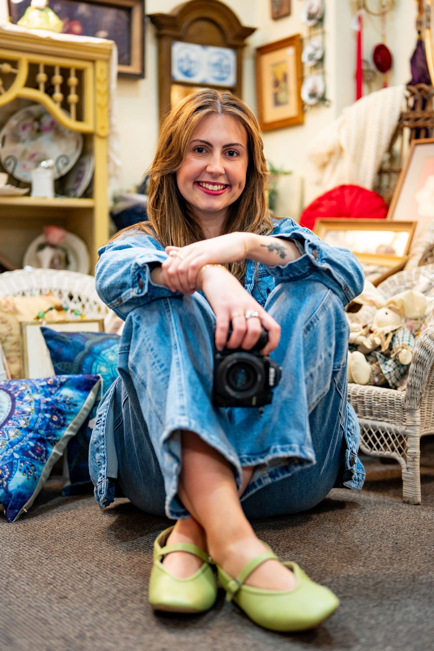 Smiling woman in denim outfit sitting on floor holding camera in vintage antique shop with decor.