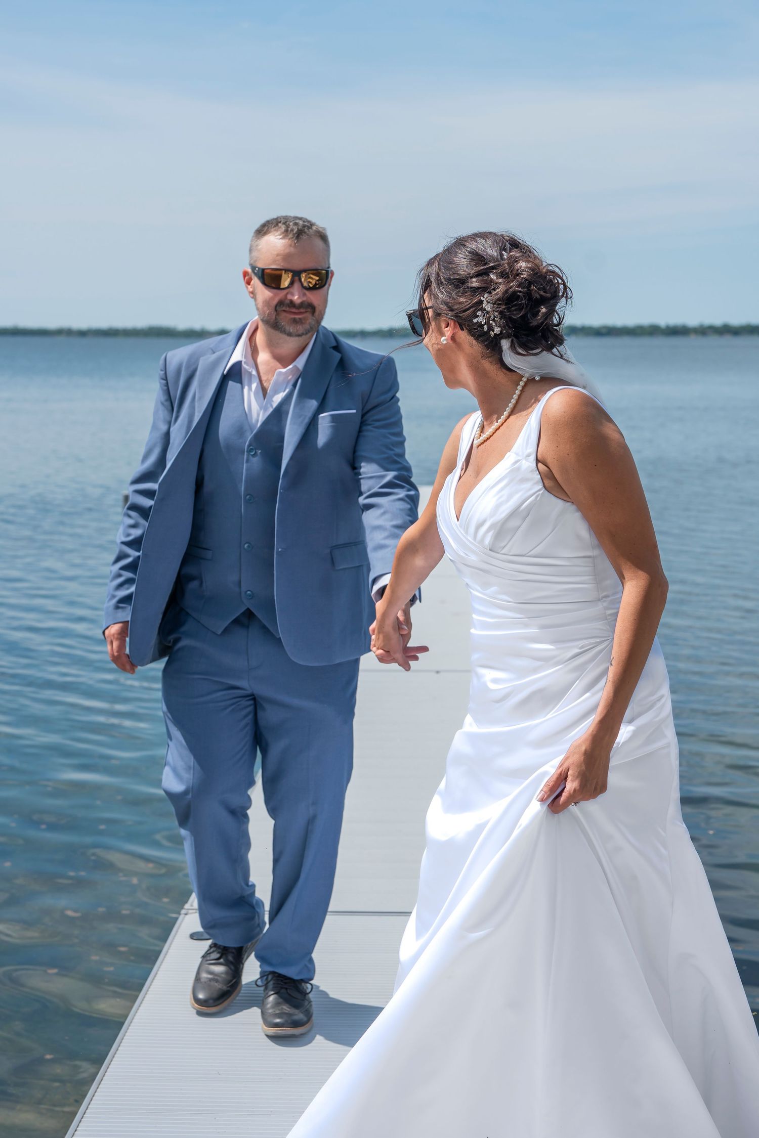Wedding couple dances and celebrates on white dock platform with lake and blue sky behind them.