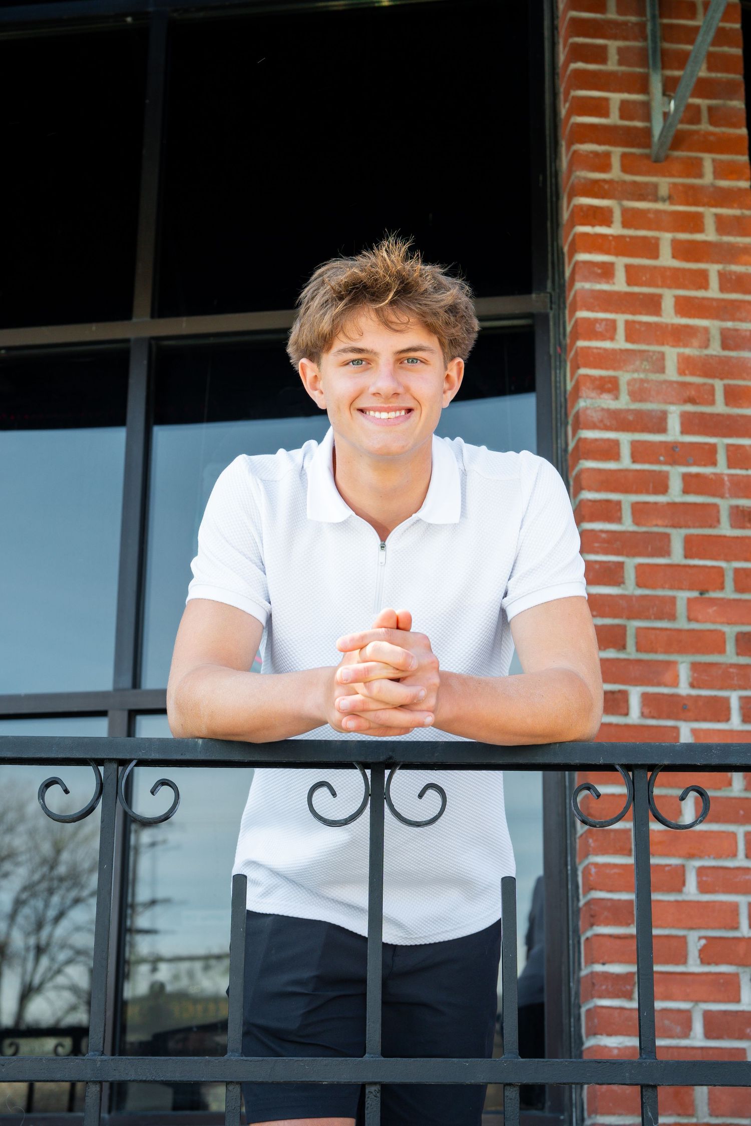 Person in a white polo shirt smiles while leaning on a black metal railing against a brick building exterior.
