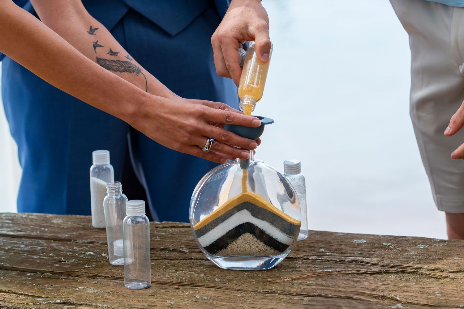 Close-up of hands pouring colored sand into decorative glass vessel during beach ceremony.