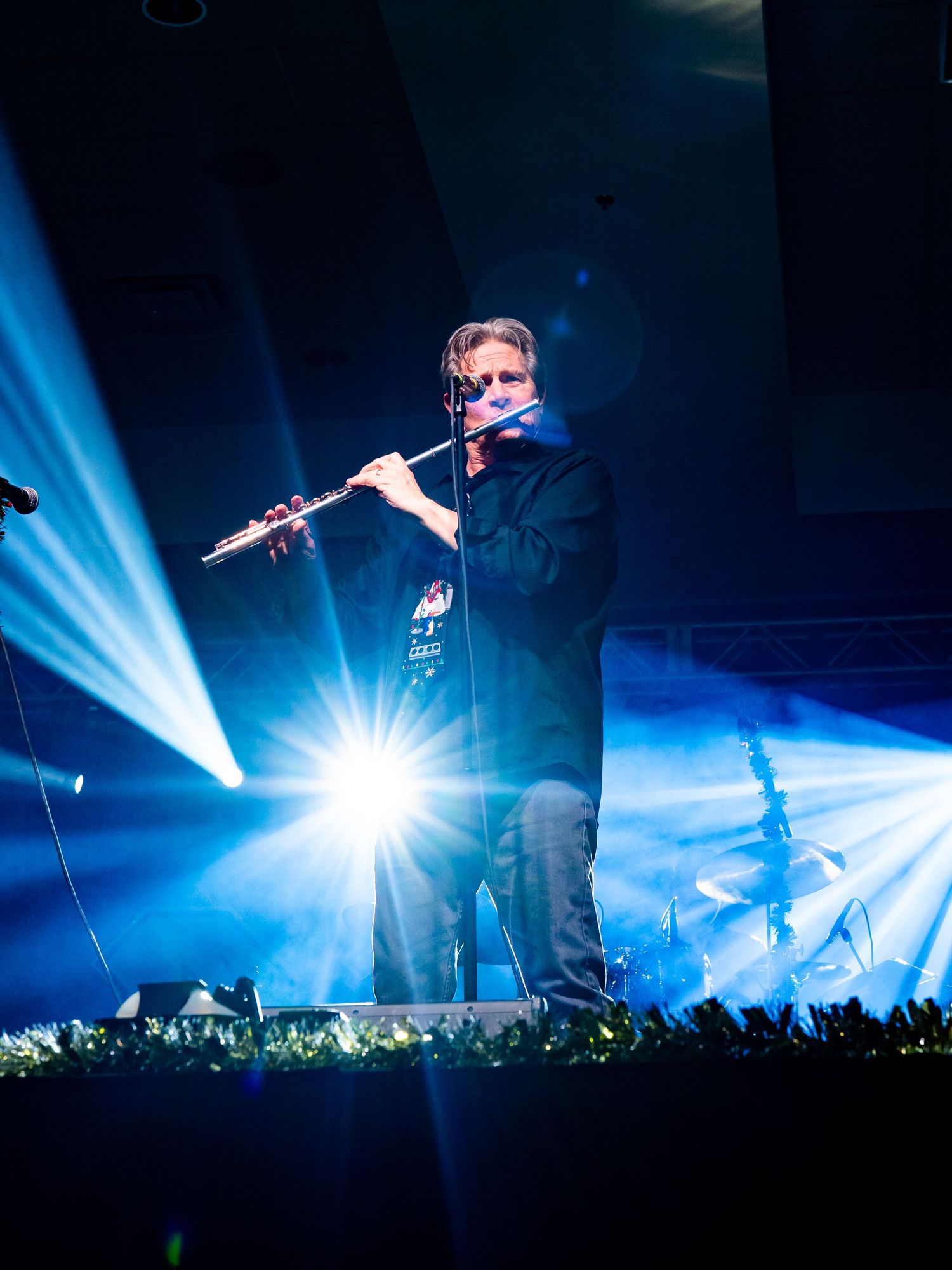 Musician performing with flute on stage illuminated by bright spotlights and blue backlighting.