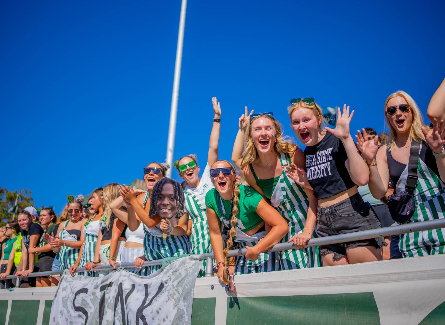 Enthusiastic crowd cheering from bleachers against bright blue sky.