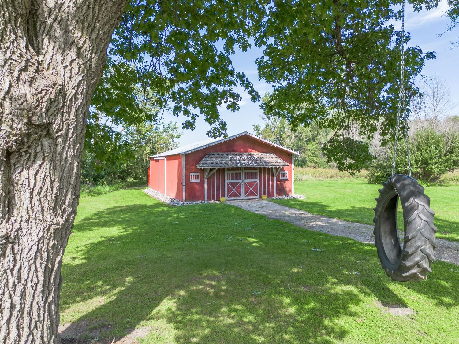 Rural property view with red barn and large tractor tires displayed along a tree-lined path.