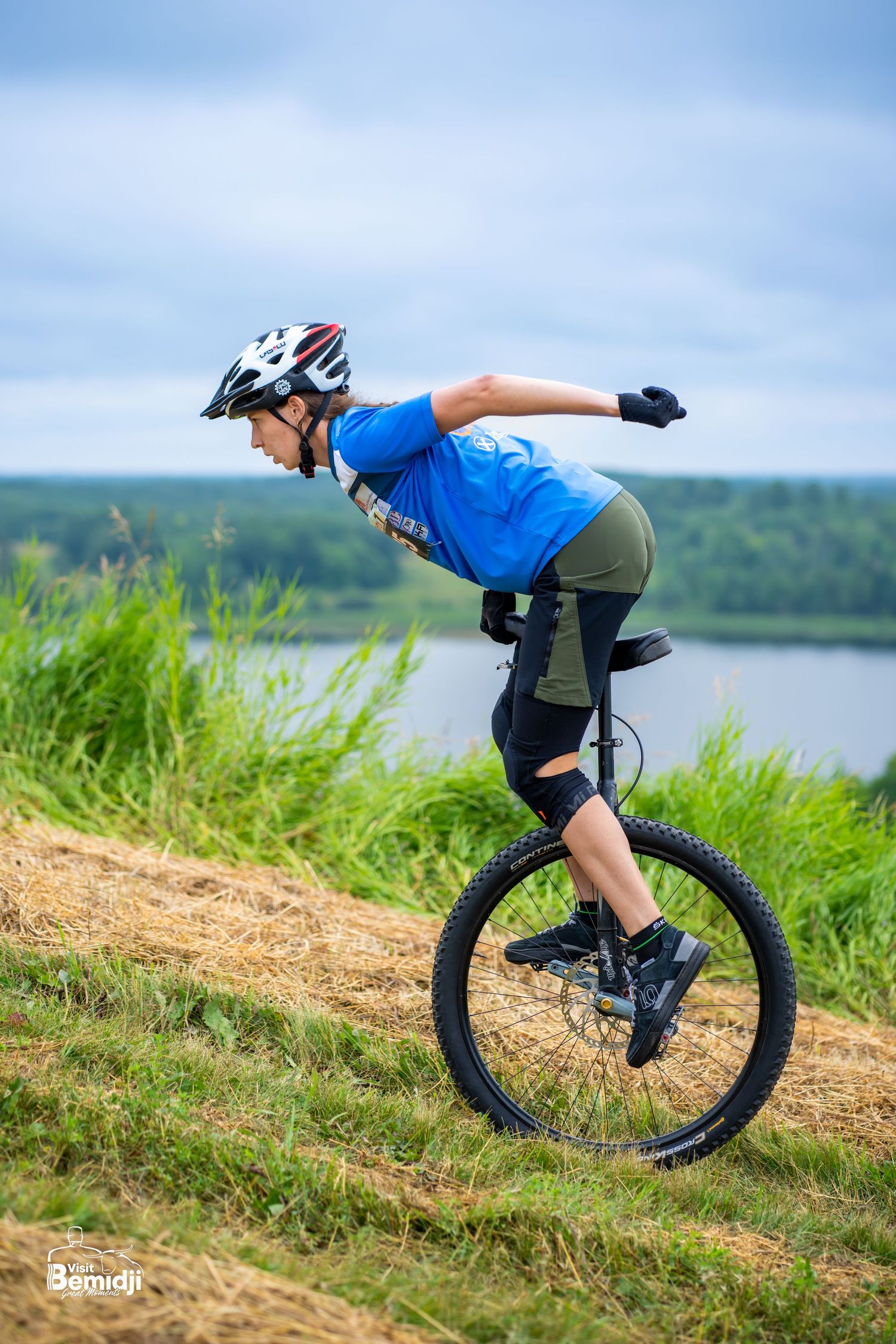Mountain unicyclist performing tricks on grassy hillside near lake.