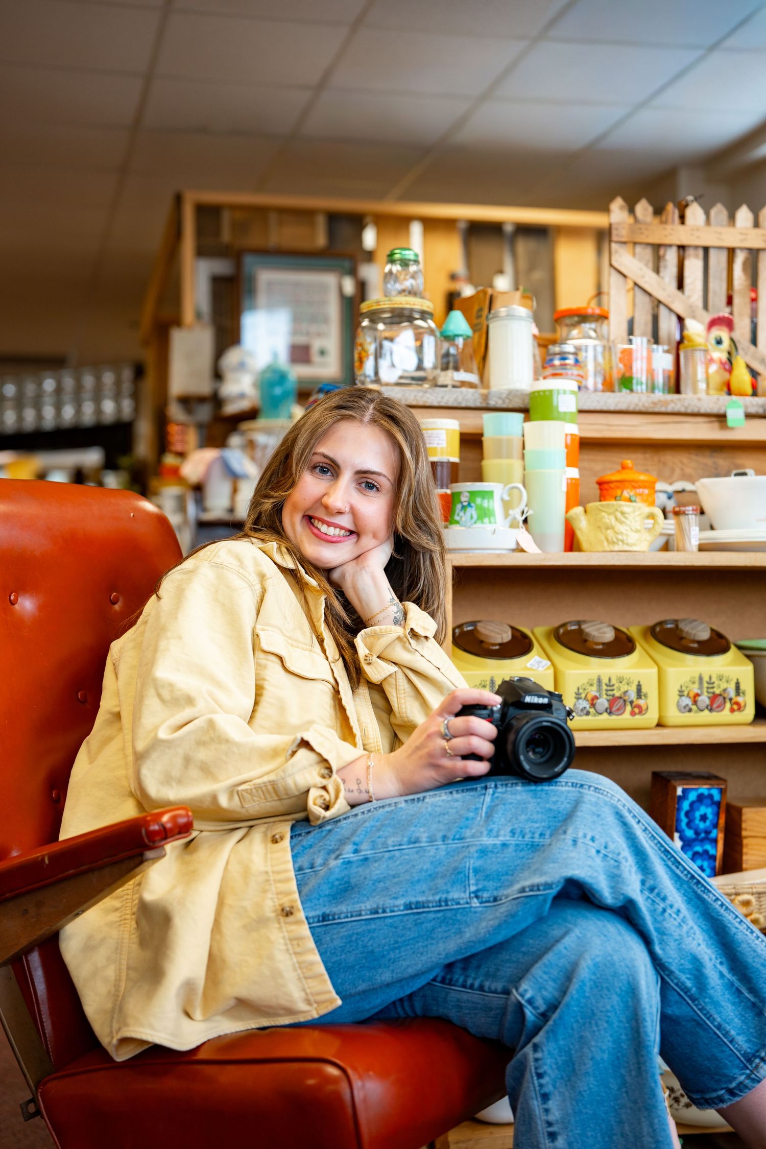Smiling woman in yellow jacket holding camera, seated in red chair at vintage shop with retro kitchenware on shelves.