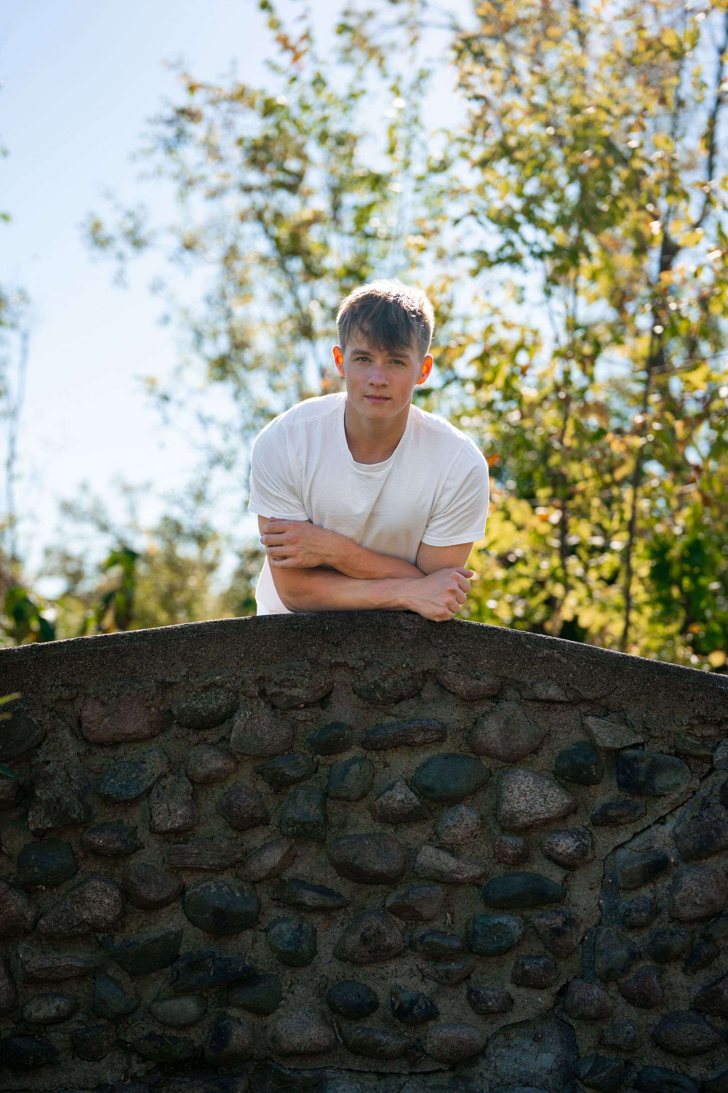Someone in white shirt leaning on stone wall with autumn trees in background.