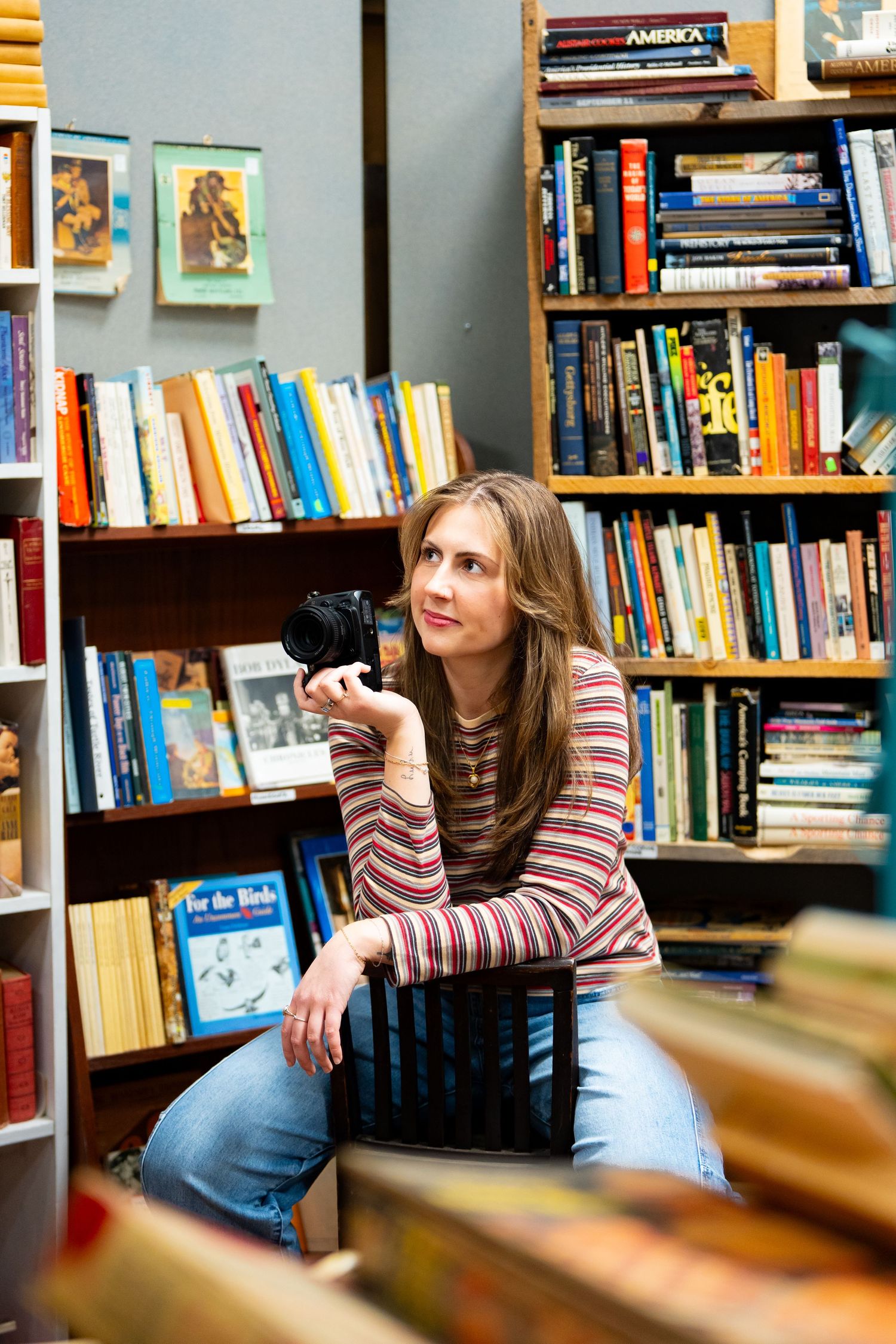Woman holding a camera sits among bookshelves in a cozy bookstore, wearing a striped top and jeans.