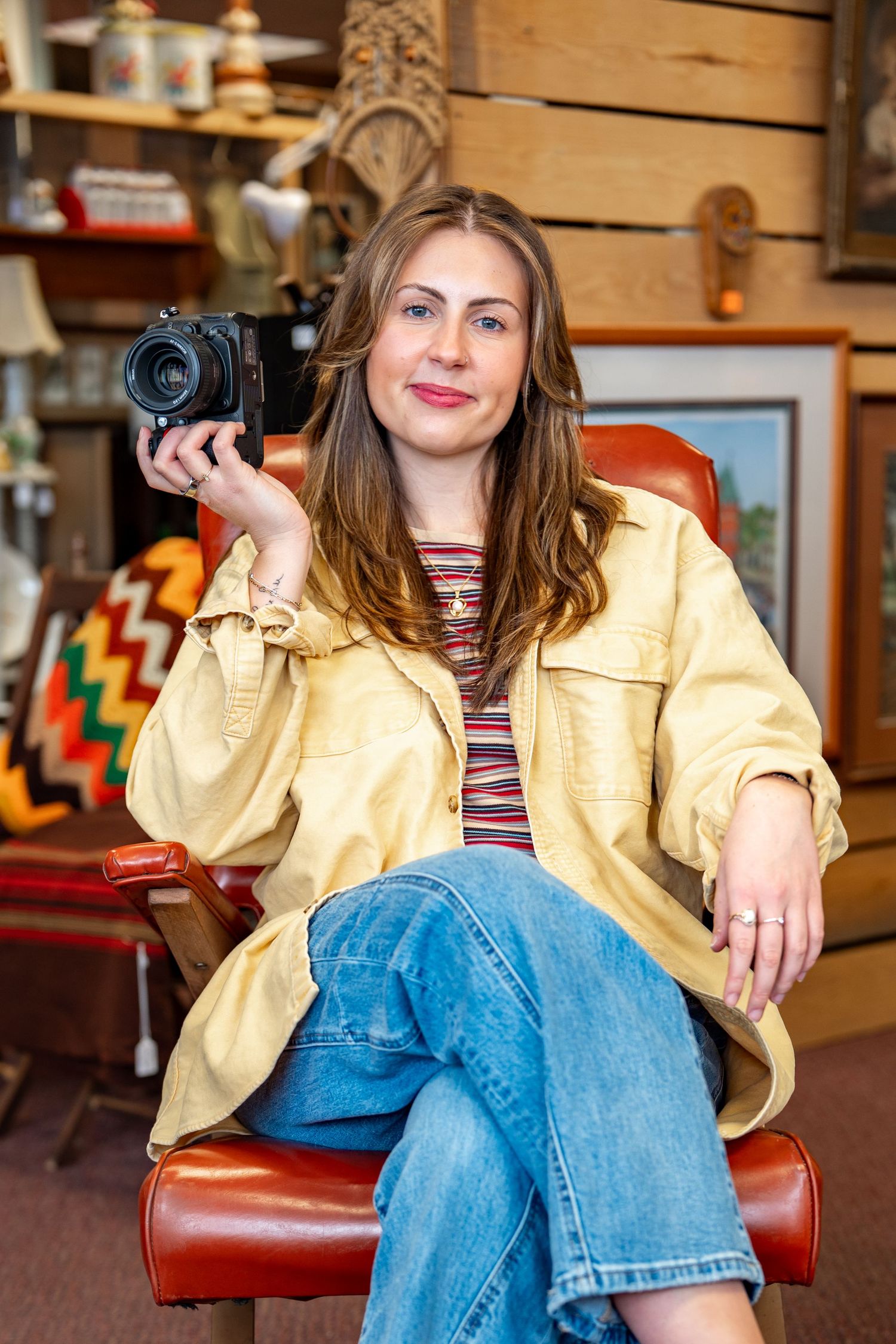 Woman in yellow jacket holding a vintage camera, seated in a red leather chair in an antique shop.