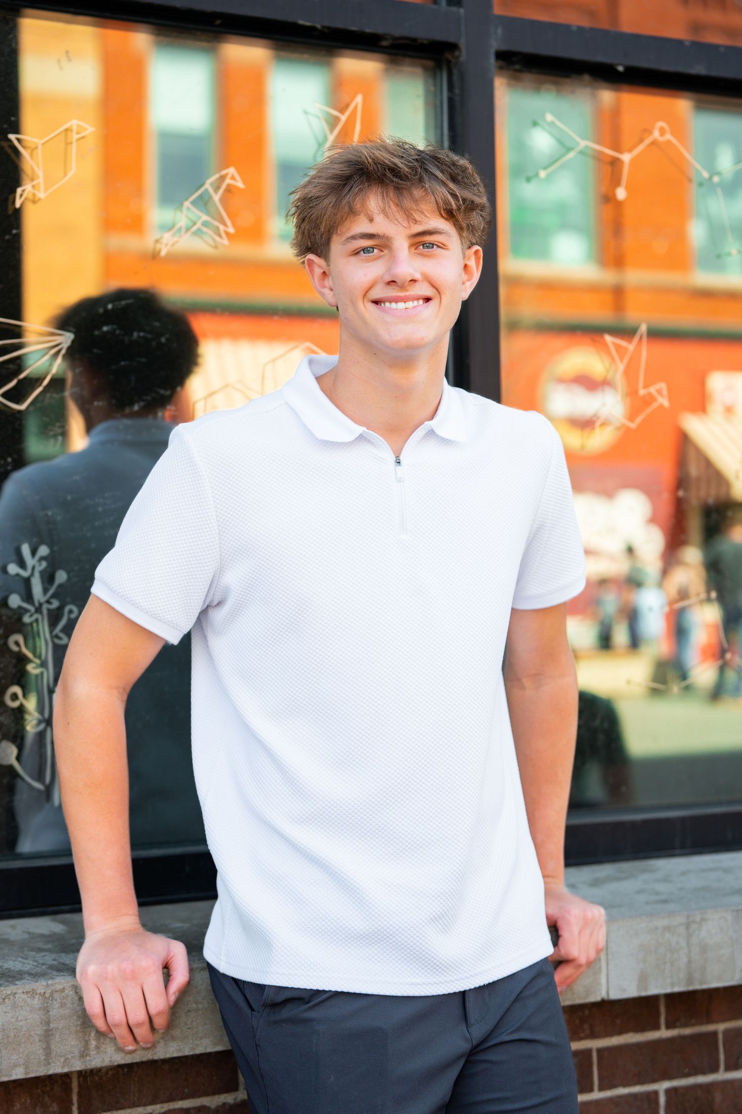 Person in white polo shirt leaning against brick wall with storefront windows in background.