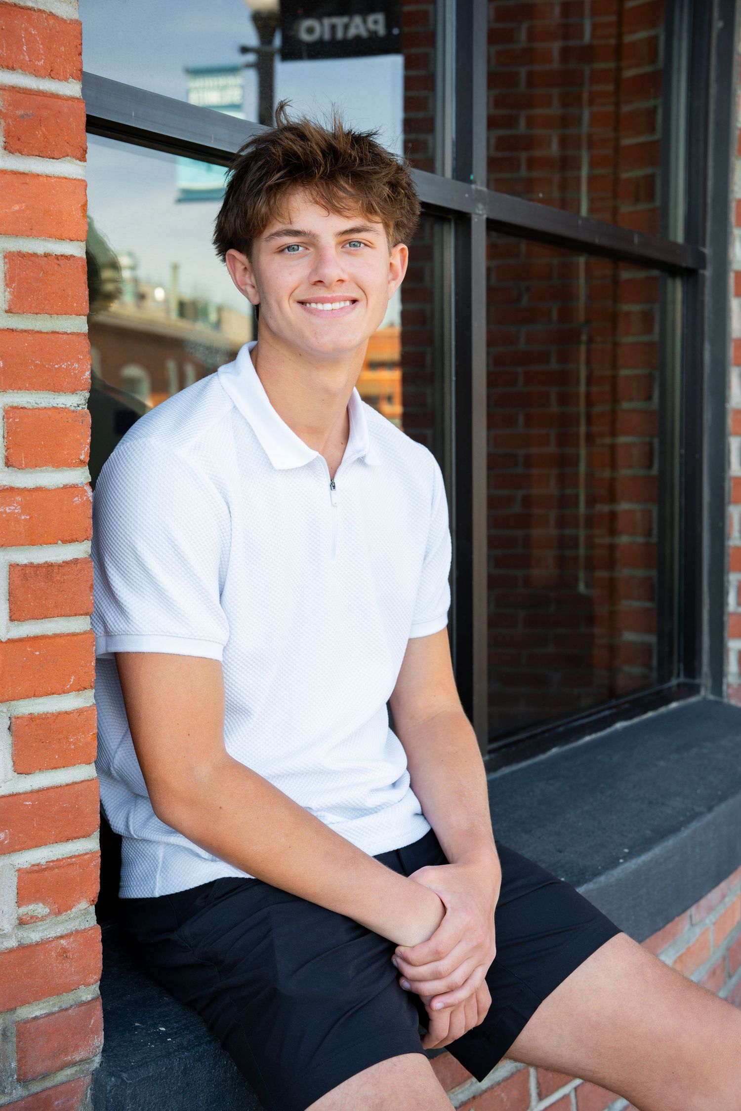 Young person in a white polo shirt and dark pants sitting casually on a brick building ledge in natural lighting.