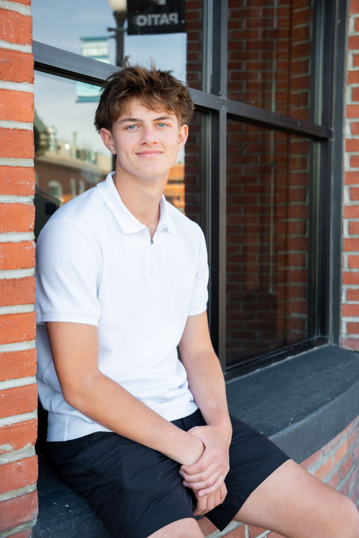 Young person in white polo shirt and dark shorts sits casually on brick ledge outside storefront window.