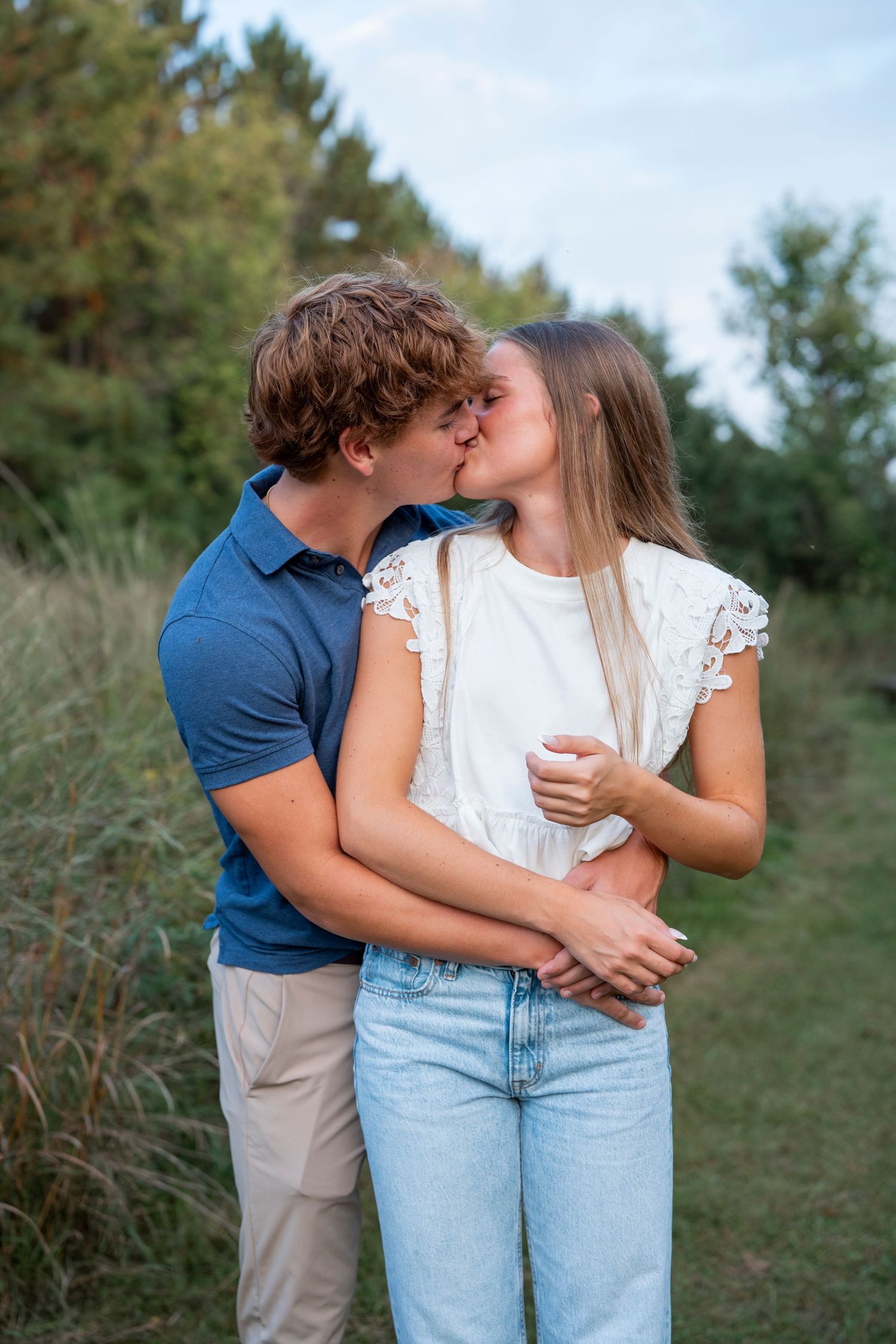 A romantic couple in casual outfits embraces on a grassy hillside.