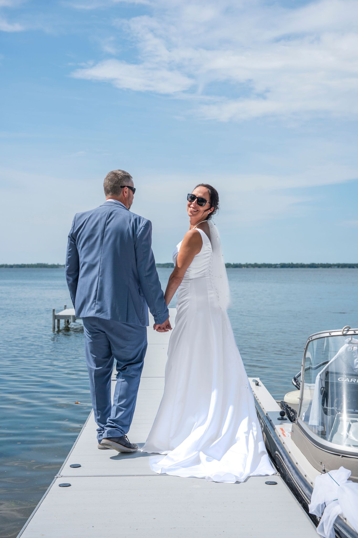 Newlyweds in blue suit and white dress share a moment on a boat dock with water and sky in background.