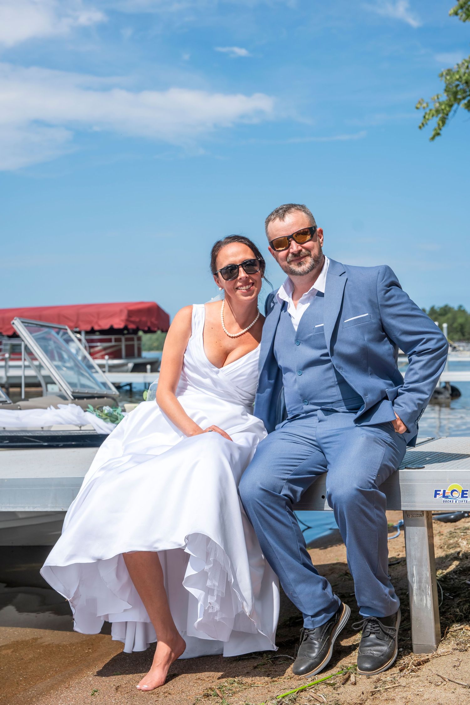 Wedding couple relaxing on wooden dock with boats and marina in background.
