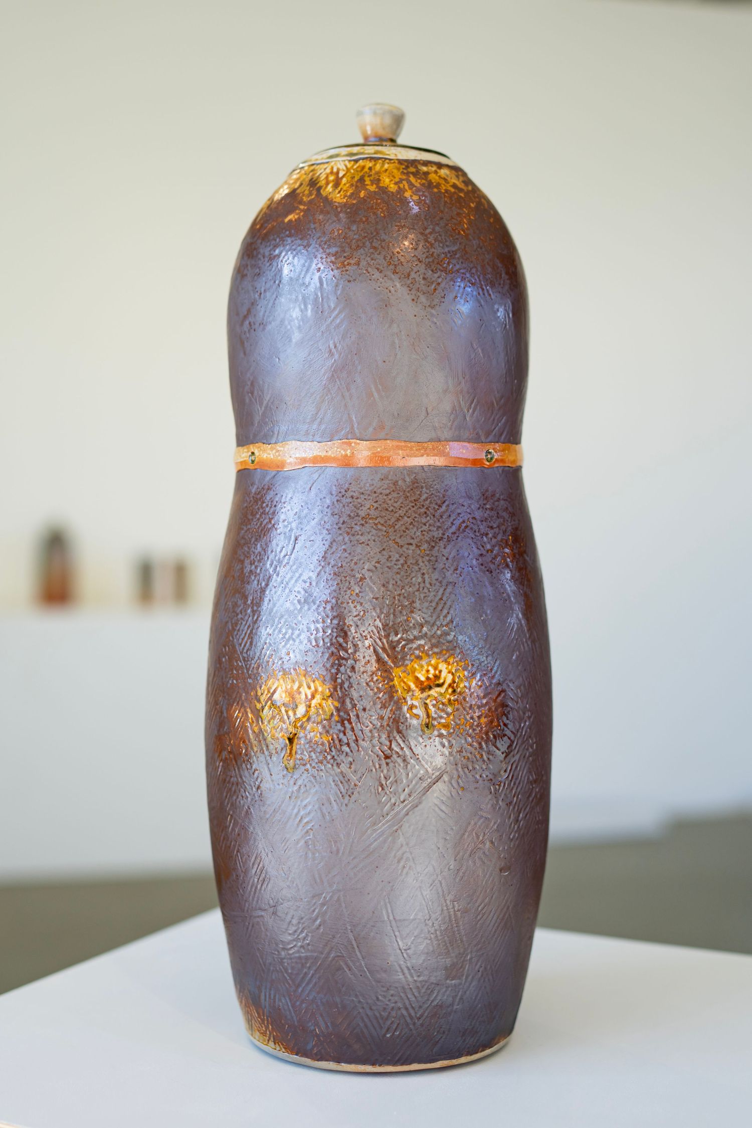 Brown ceramic vessel with metallic copper band detail displayed on white surface at art show.