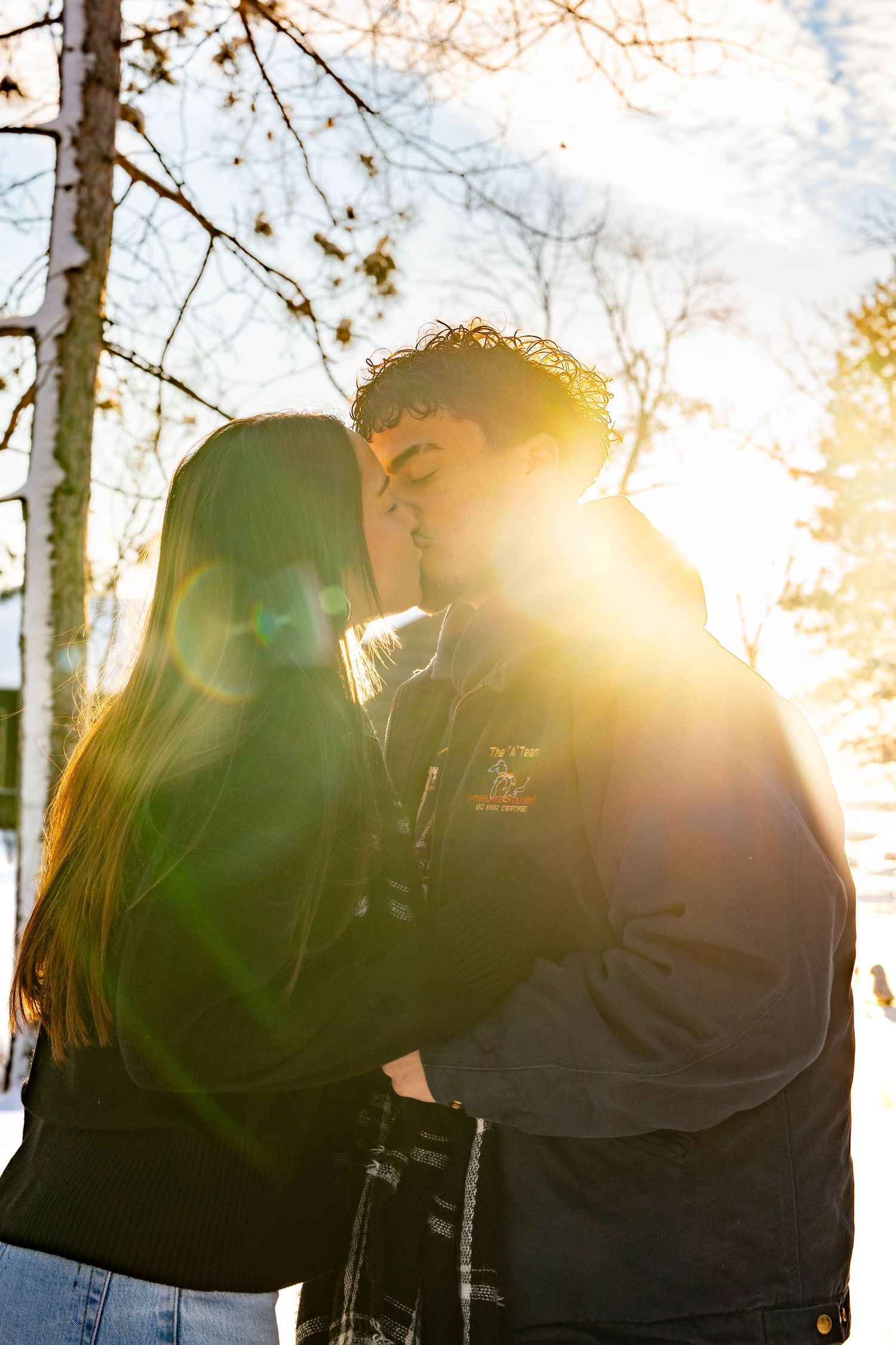 A silhouetted couple stands together in a forest with bright sunlight streaming through trees creating a warm lens flare.