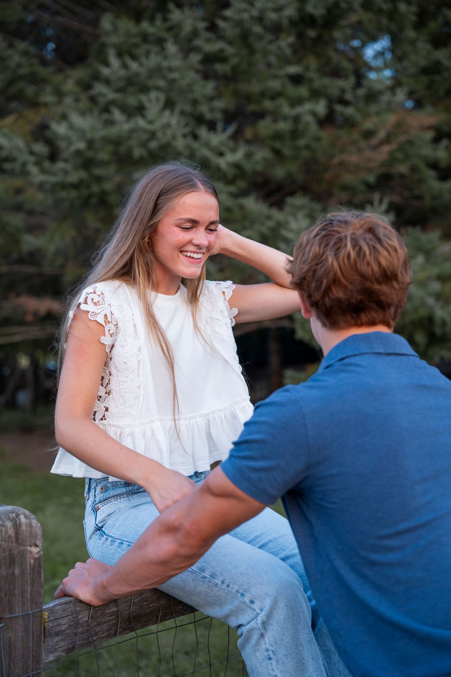 A playful outdoor photoshoot scene showing a couple sitting on a wooden fence surrounded by trees.