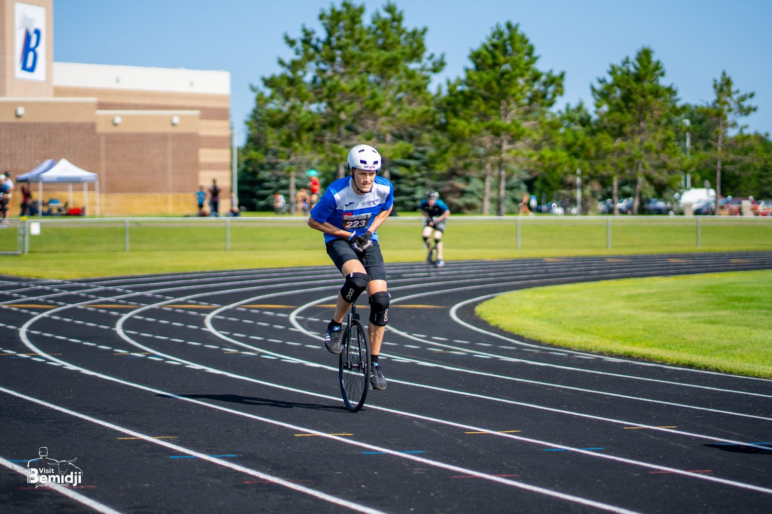 Solo unicyclist performs on blue running track with trees in background.